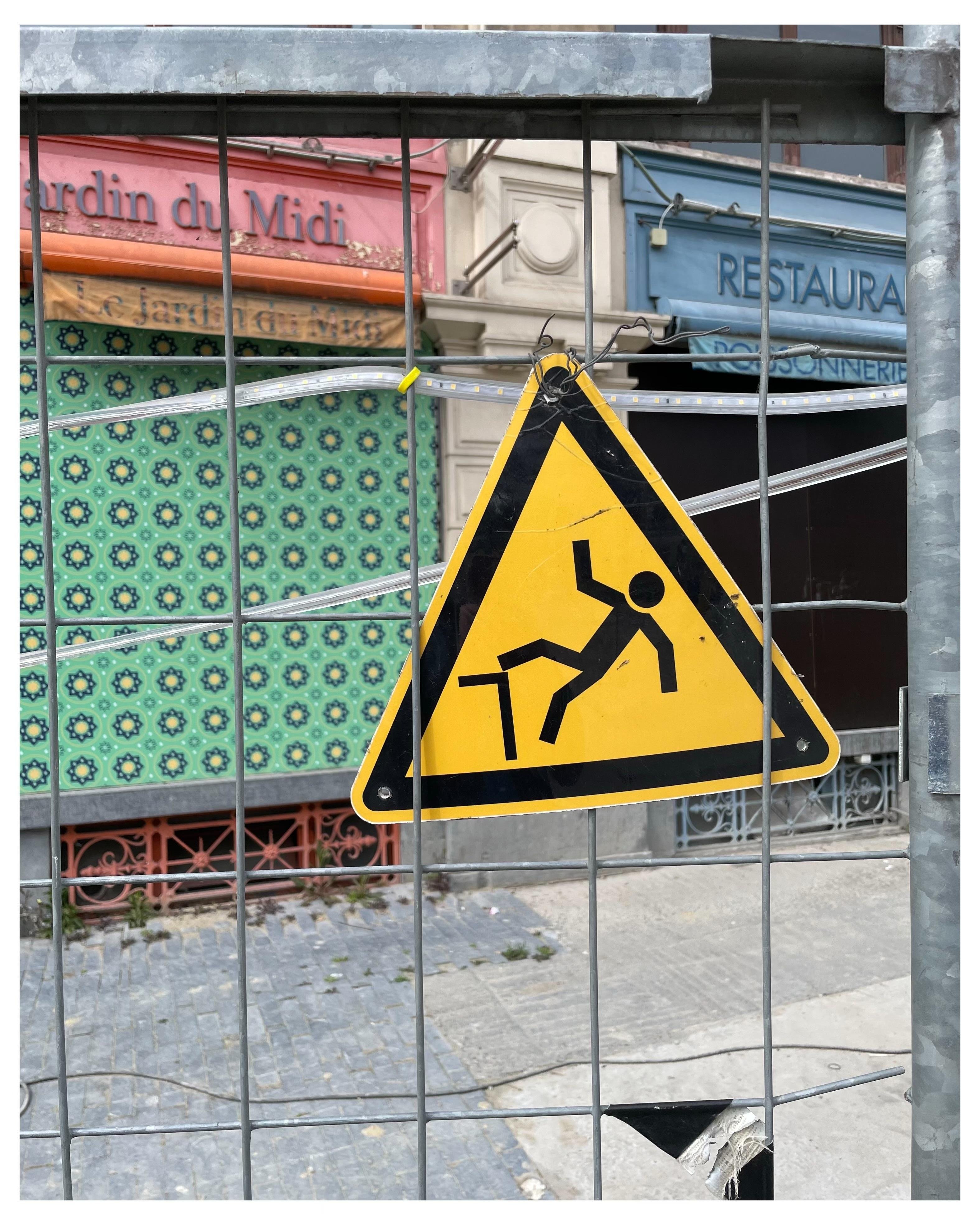 Construction site, metal wire fence with yellow caution triangle depicting a stick man falling backwards off a tall step or ledge.