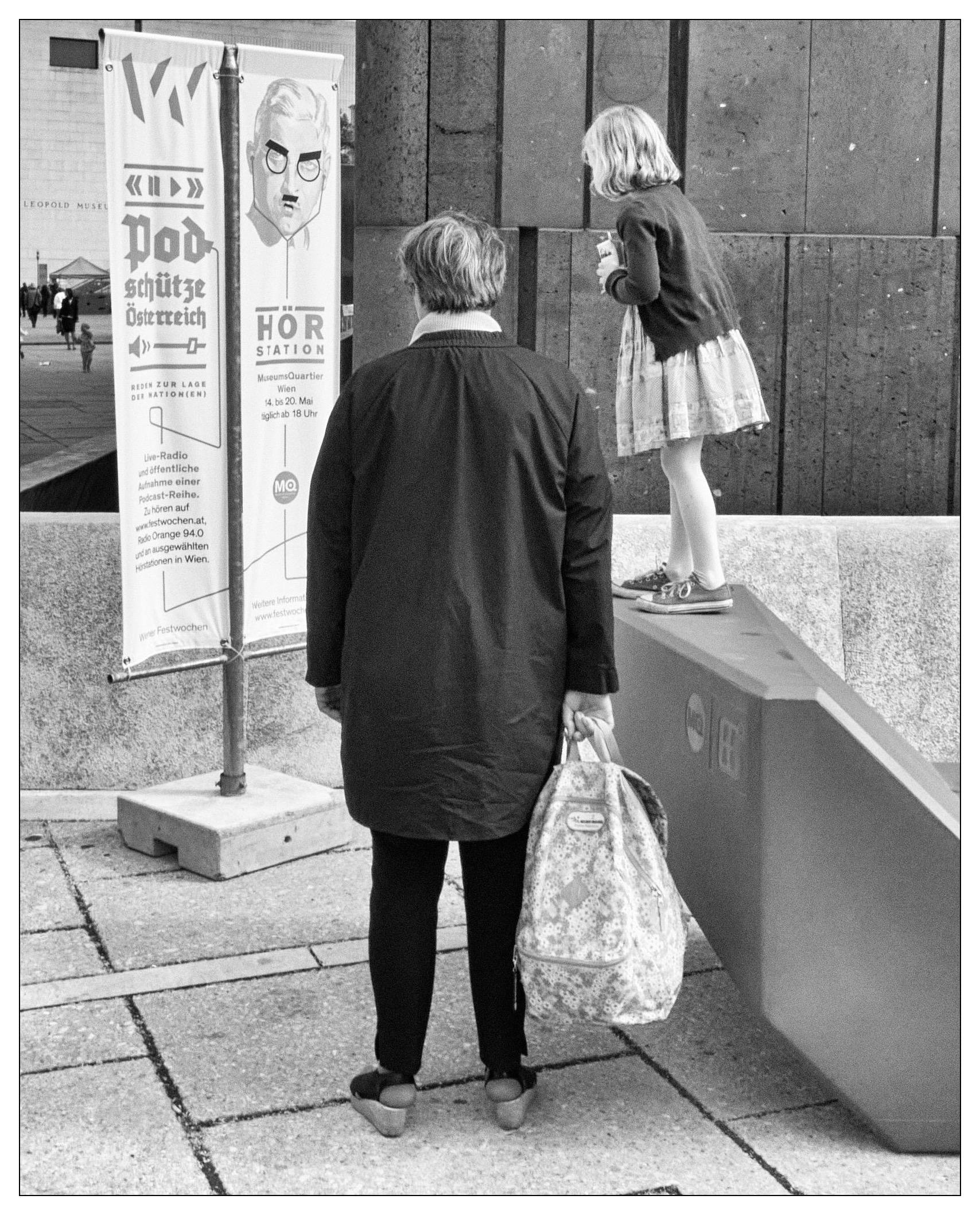 This black and white photograph displays a touching scene featuring two individuals and a cultural setting. On the right, a young girl stands atop a raised concrete ledge, looking at a vertical banner. Opposite her, an older adult woman stands facing the girl and the information banner. This adult is dressed in a black coat and slacks, complementing the simplicity and elegant ambiance of the scene. They hold a patterned bag and appear to be either watching the girl or engaging in conversation with her.

To the left of the image, there’s a banner featuring a graphic design and text related to an event or exhibition, indicative of a cultural or possibly museum district. The surroundings, including visible architectural elements such as concrete walls, suggest a public, urban space.

Overall, the image conveys a sense of casual public interaction, possibly within a cultural or educational setting, emphasizing a moment of simple human connection.