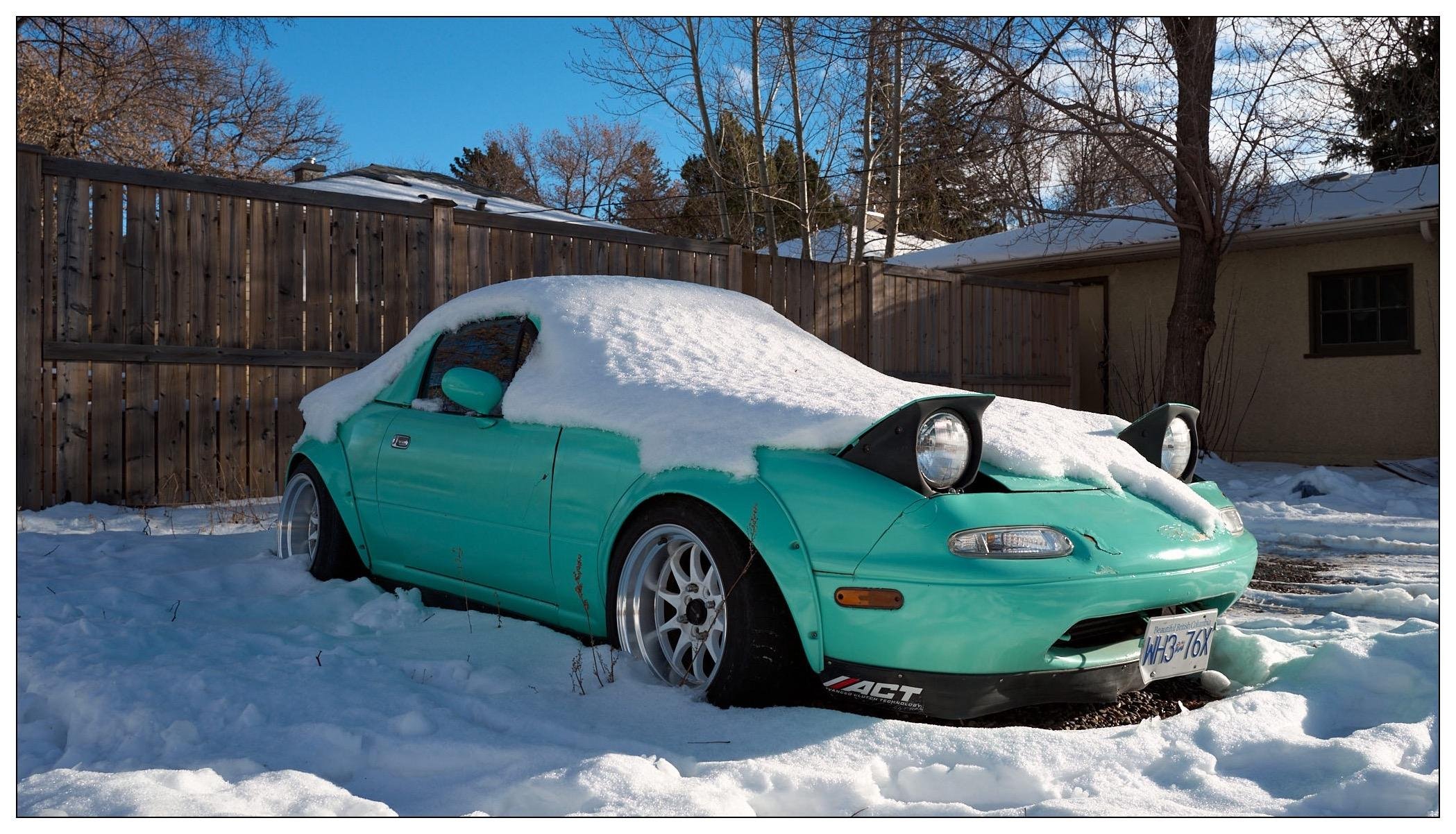 A teal mazda miata sports car partially covered in snow is parked in a residential area next to a wooden fence. The ground is covered with snow and patches of grass peek through. Bright sunlight illuminates the scene, casting shadows on the snow. The car's headlights are pop-up style, and it features a cracked front bumper and white wheels.