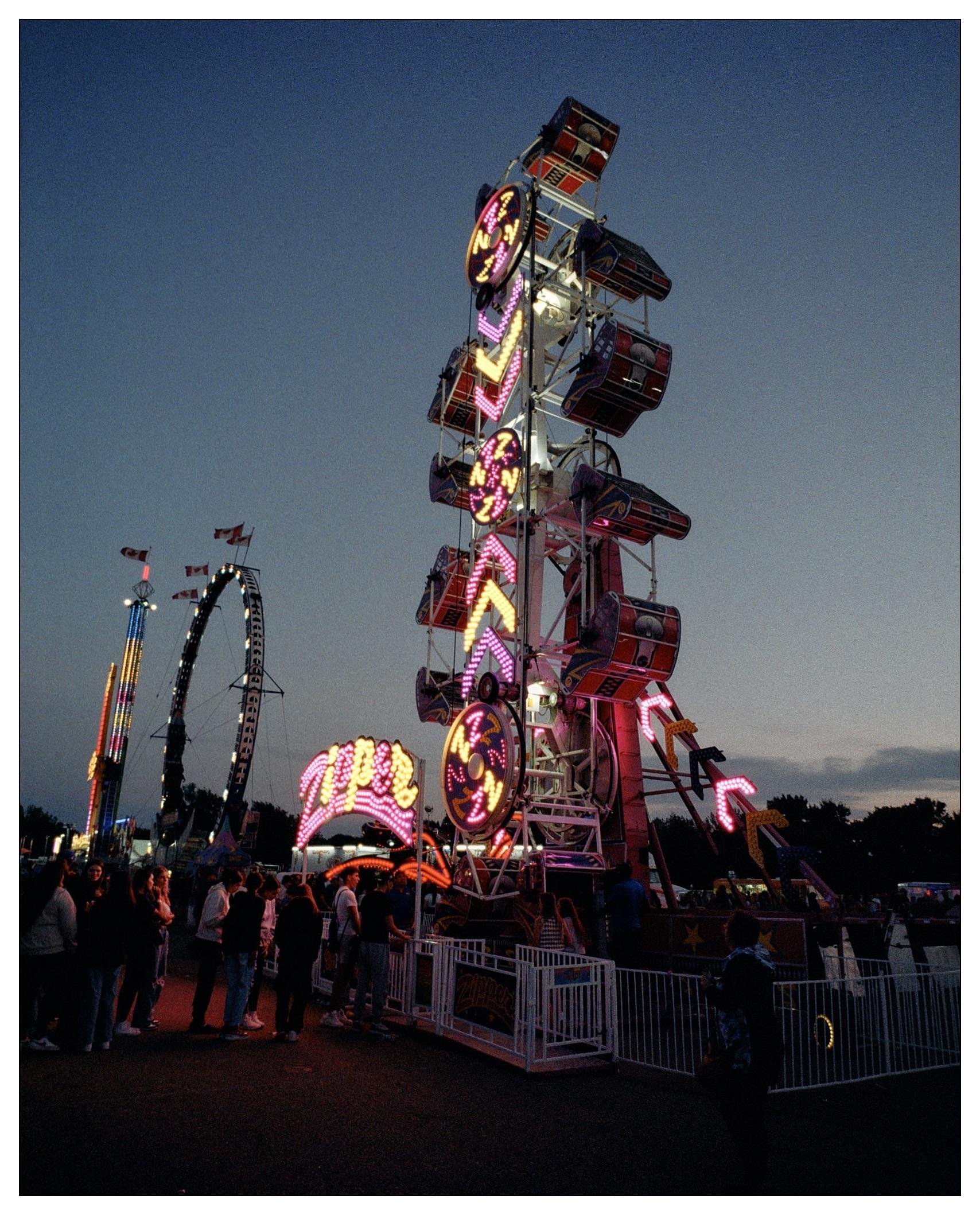 This image shows a vibrant scene from a carnival or amusement park at dusk. It features a large, brightly lit ride called the Zipper, packed with visitors. The frame captures the motion and lights of the ride, and the atmosphere is lively with a fairly large crowd of people milling about, enjoying the evening. There are also other amusement rides visible in the background, including what appears to be a part of a roller coaster and another attraction adorned with lights. The sky is dimly lit, indicating it is either early evening or nighttime.