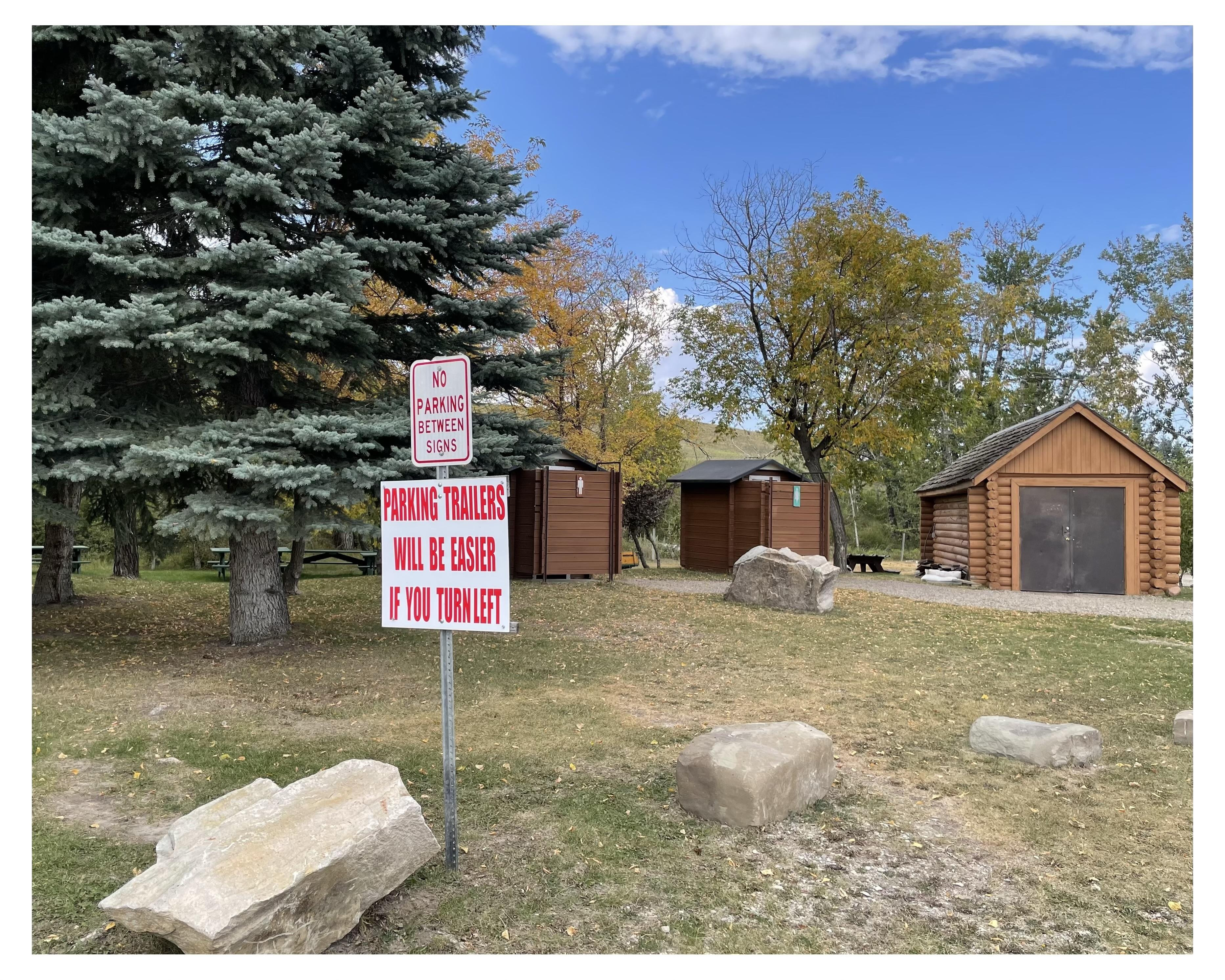 Two red and white signs mounted on one post in a park setting, with the top sign reading "NO PARKING BETWEEN SIGNS" and the bottom sign advising "PARKING TRAILERS WILL BE EASIER IF YOU TURN LEFT". In the background, there are two wooden cabins and a large evergreen tree to the left. Some large rocks and patches of grass are visible in the foreground.