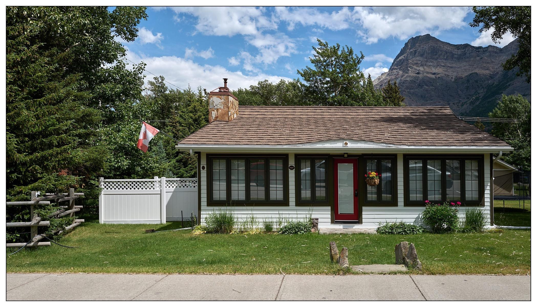 Small single-story house with a white fence, located in a rural setting with a mountain in the background. The house features a red door and green trim around windows, a hanging flower basket on the door, and a Canadian flag flying to the left. Nearby, there's a weathered wood post fence partially visible, with mature trees and shrubs around the property.