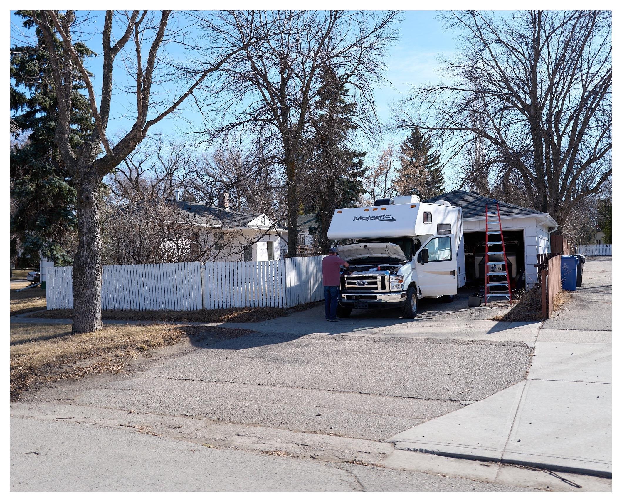 A residential area featuring a large RV parked in a driveway next to a garage with a white picket fence. There are trees around the area, indicating a suburban setting. The RV prominently displays a "Majestic" logo on its over-cab section. Additionally, there is a person visible, standing beside the RV, possibly performing some maintenance tasks as they have the hood open and also nearby is a ladder. The scene is characterized by a clear sky and a tranquil suburban vibe, with a road in the foreground and residential houses in the background.