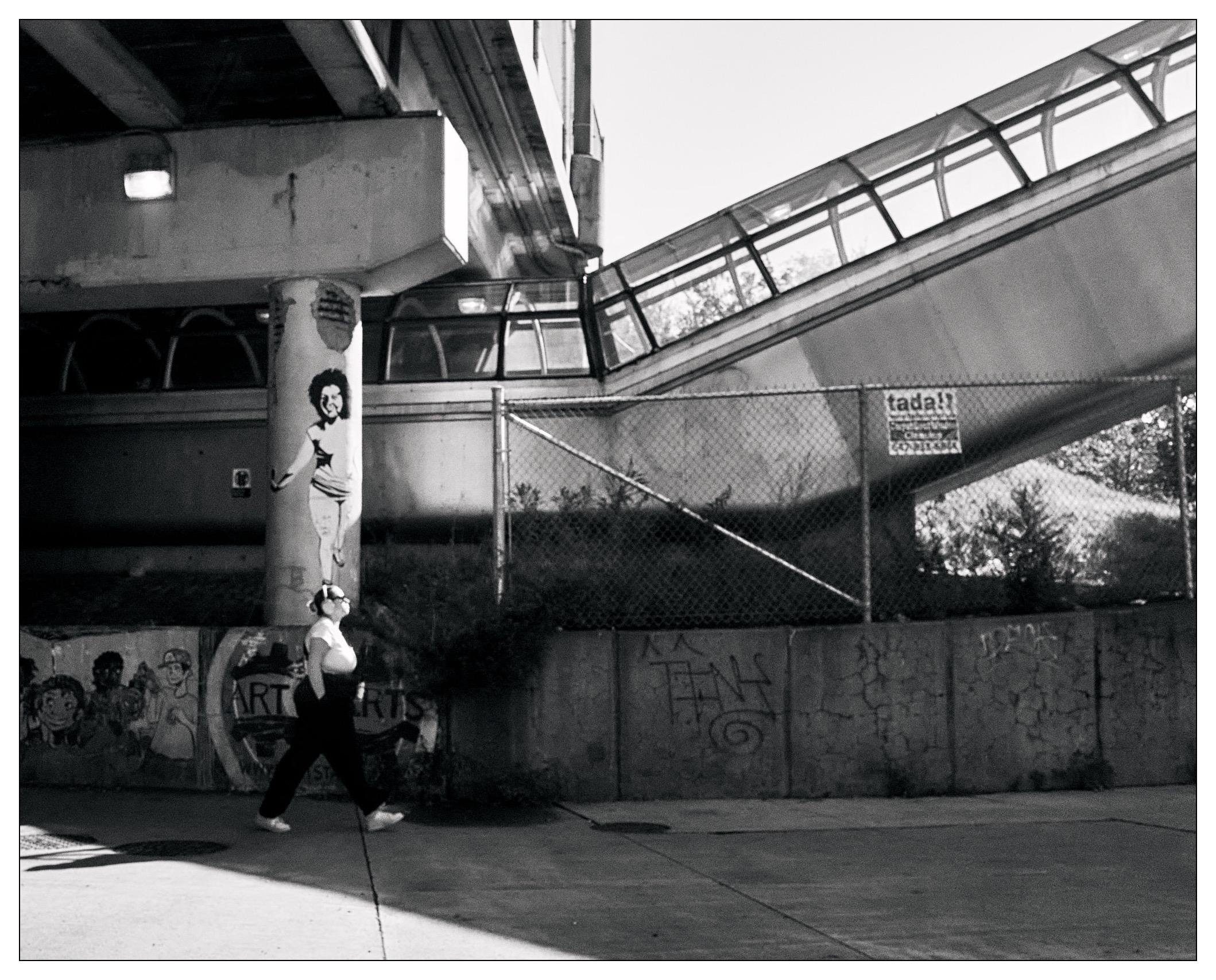 Black and white photograph of an urban scene featuring a person wearing over the ear headphones, walking past a graffiti-covered column and concrete walls underneath a bridge complex with elevated walkways and ramps. The column is adorned with a poster of a smiling woman posing playfully. The poster is aligned with the person walking, making it appear that the woman in the poster is balancing on the walker's head. There are various graffiti tags and drawings, including caricatures and the word "ART" on the surrounding walls.