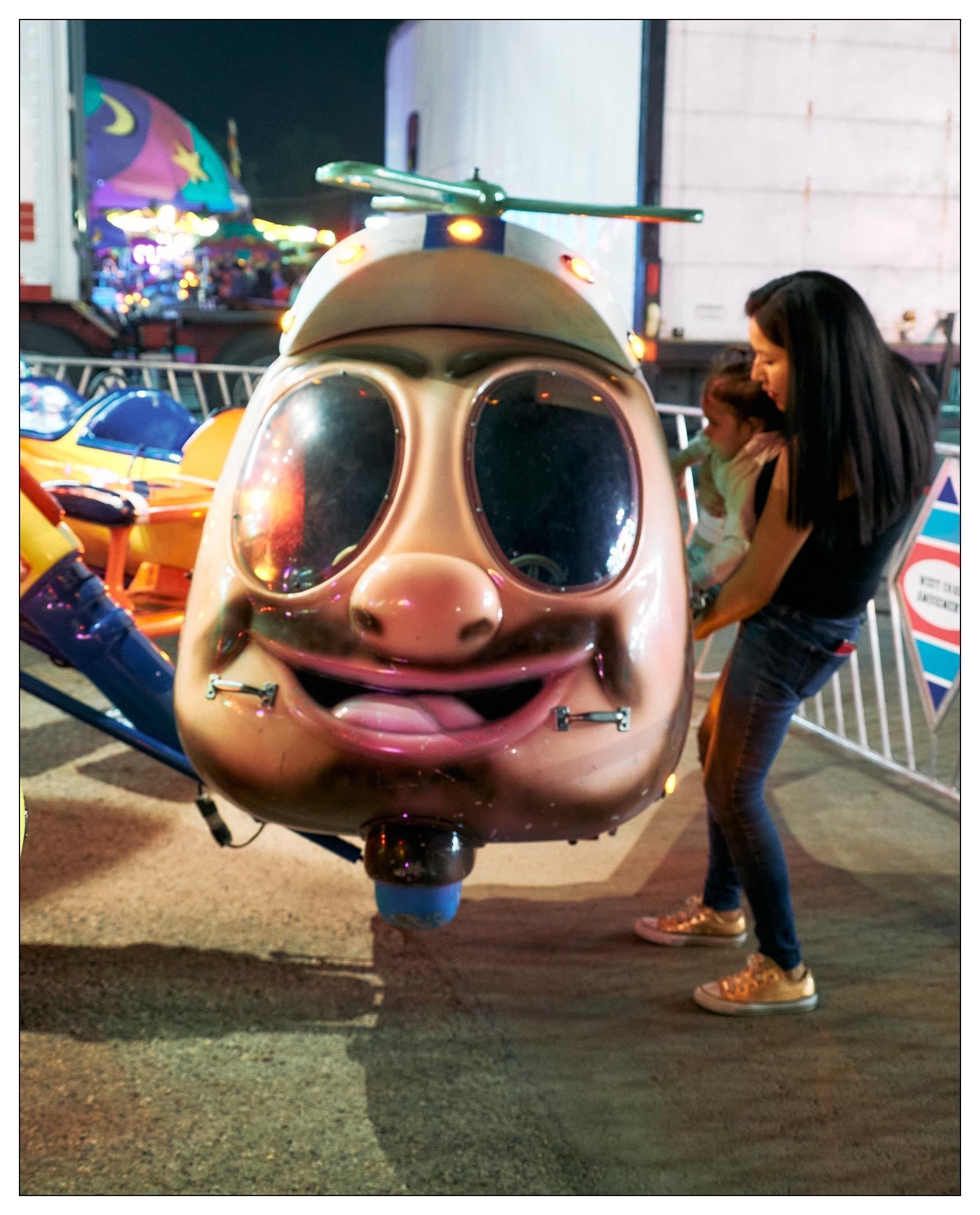This image captures a fun scene at what appears to be a fair or amusement park. In the foreground, there is a whimsical helicopter ride designed to look like a cartoonish man with a big, friendly smile, large sunglasses, and helicopter blades on top. A woman is standing next to it, helping a small child who is getting into the helicopter. In the background, you can see colorful lights and parts of other amusement park rides which contribute to the lively and festive atmosphere of the setting.