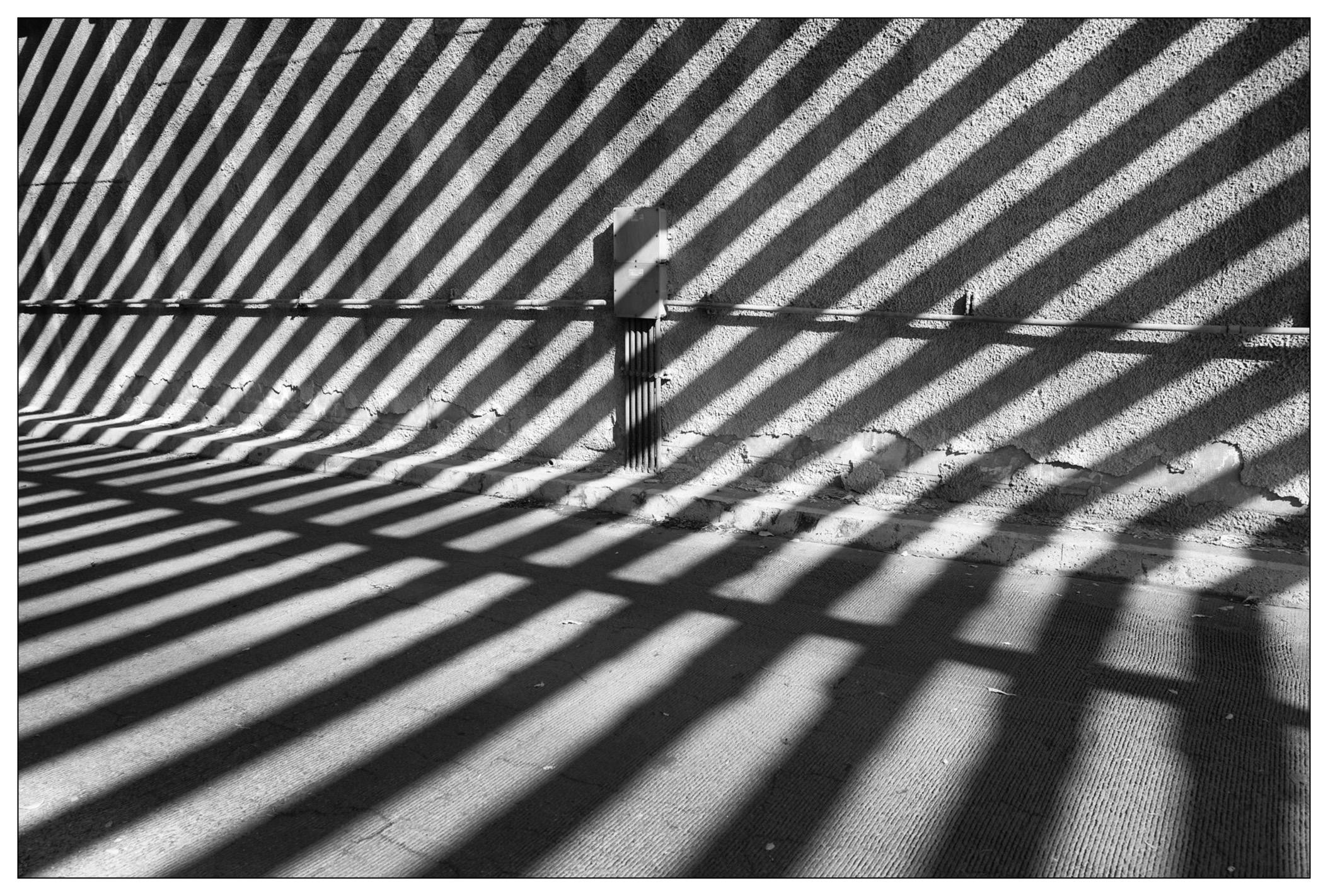 Black and white photo of strong diagonal shadows cast by a fence on a textured ground with a singular utility box and conduit centrally positioned, creating contrast between light and shadow.