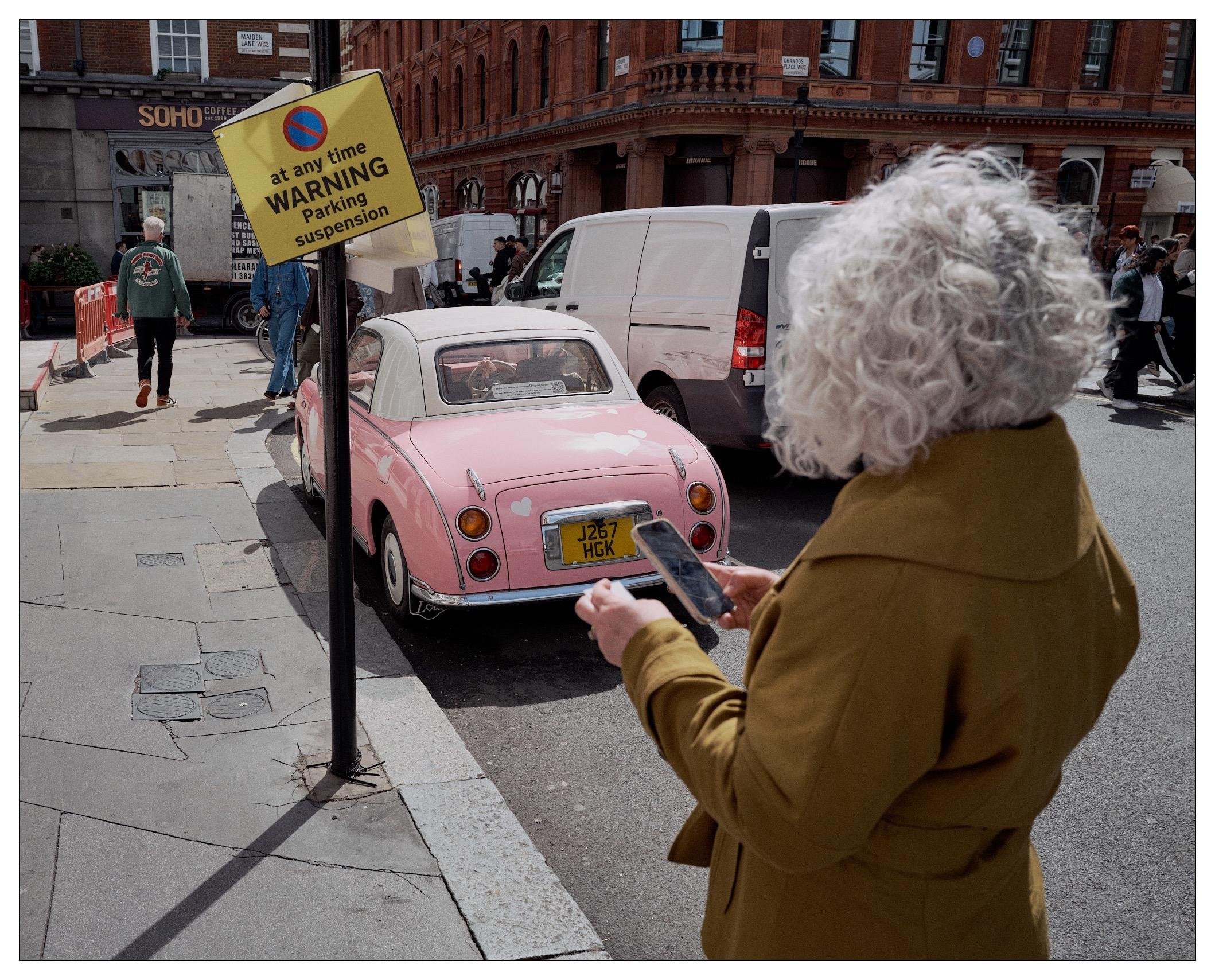 Street scene, small pink classic car with white roof parked next to a no temporary no parking sign.  Woman with striking curly white hair and a tan overcoat stands in front contemplating taking a photo with her mobil phone.