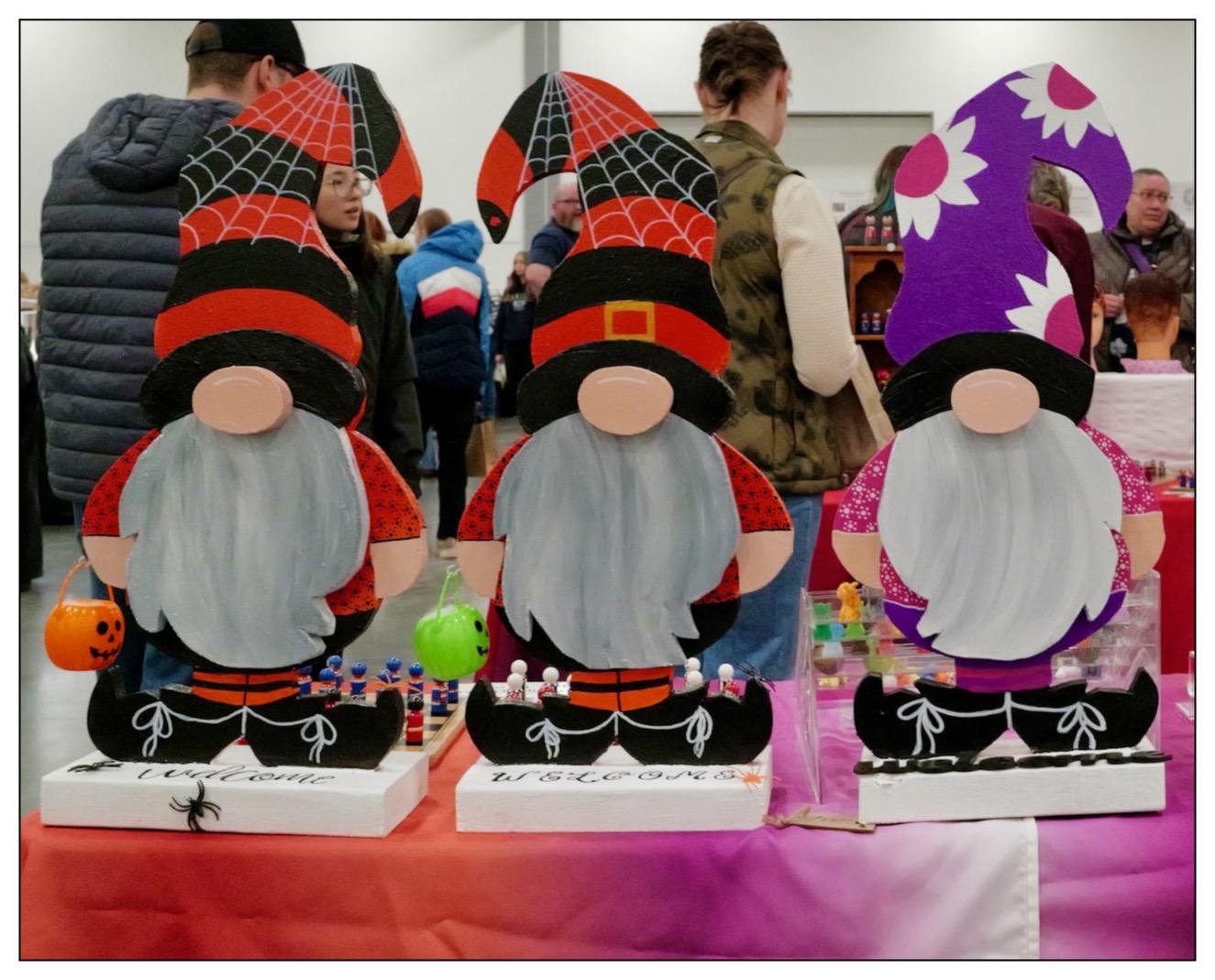 Three decorative gnome figures displayed on a table at a crafts fair. From left to right: the first gnome wears a tall black and red spiderweb-patterned hat, an orange and black outfit, and carries a small orange pumpkin bucket. The second gnome's outfit and hat are similar to the first, except the hat is entirely red-patterned. The third gnome has a tall, curling purple hat with flower patterns, and a sparkly pink top. Each gnome is bearded, with a white, flowing beard that covers its lower face. Miniature Halloween-themed decorations, including spiders and candies, are arranged in front of each gnome. In the background, attendees wander the fair.