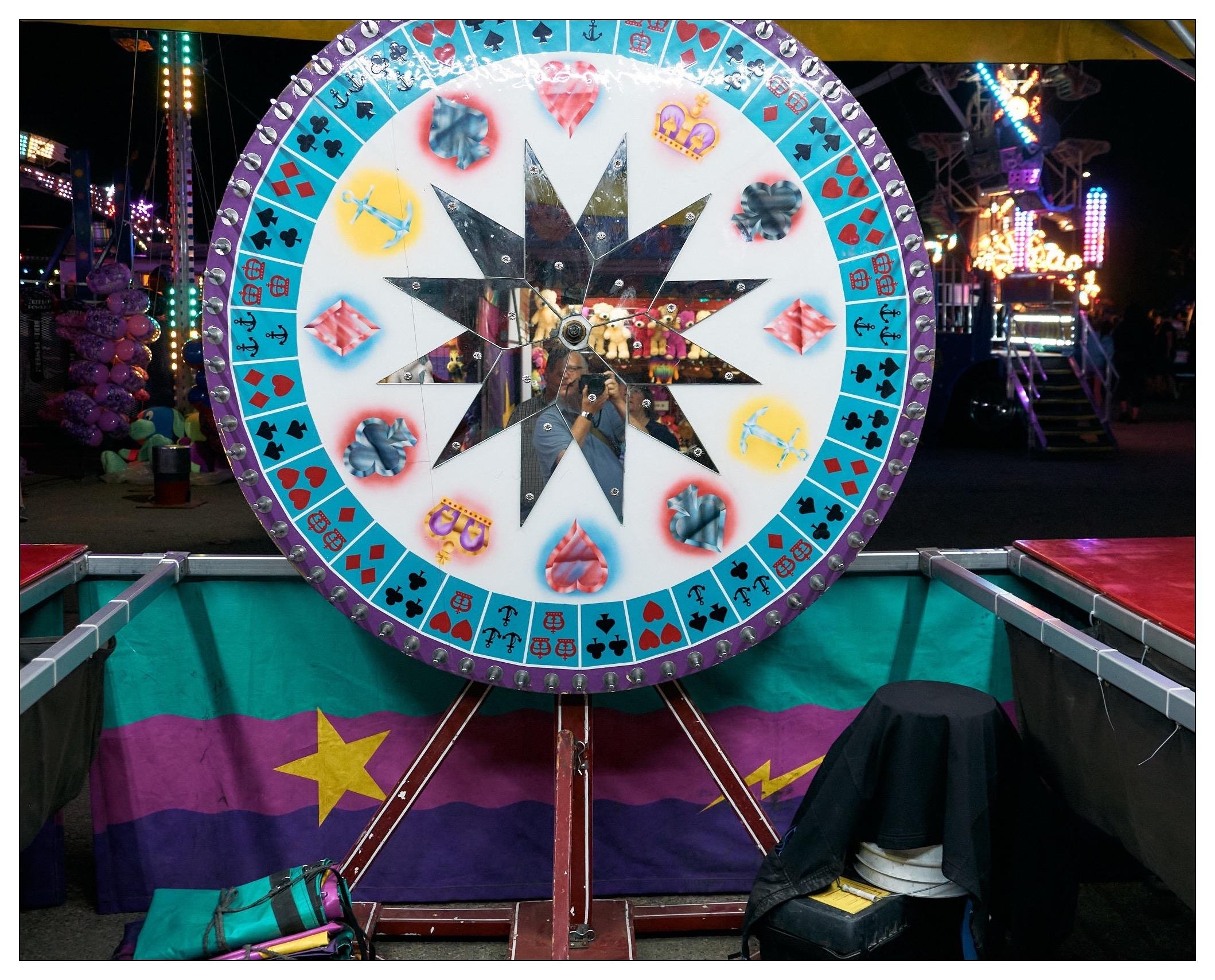 The image shows a brightly colored game at a fair or amusement park. It features a large wheel with a star design in the center and various colorful patterns and symbols on the outer rim. Visible in the background are carnival lights and other attractions, adding to the festive and vibrant atmosphere typical of such events.