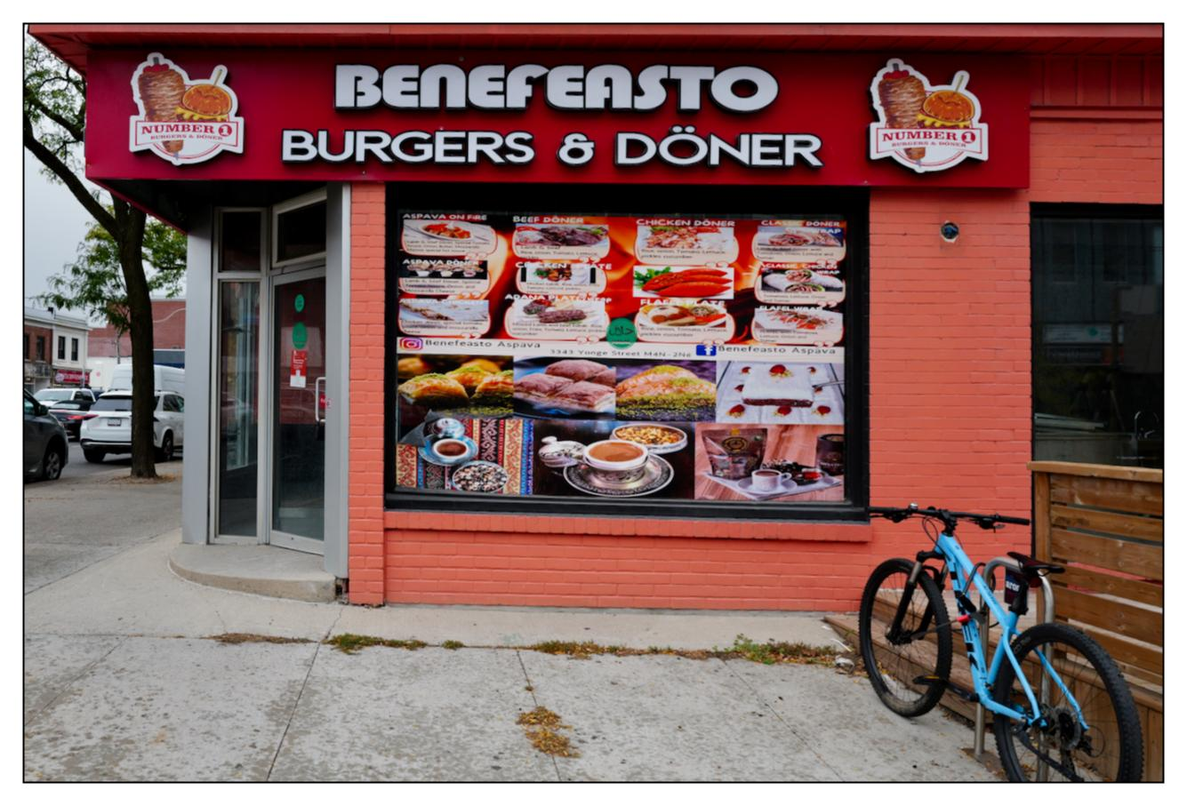 Corner view of Benefeasto Burgers & Döner restaurant with a bright red facade featuring a large menu display and restaurant signage in both English and unidentified non-Latin script. A blue bicycle is parked on the sidewalk in front of the restaurant. Signs and parking are visible in the background.