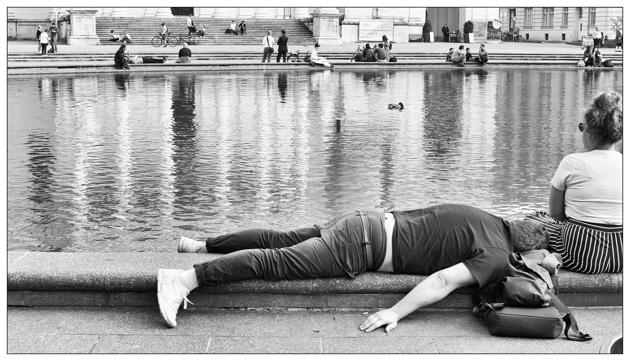 The image portrays an urban scene by a large reflecting pool. In the foreground, a person is lying face down across a ledge at the edge of the pool, seemingly relaxed or perhaps fatigued, with a backpack resting beside them. On the other side, another individual is seated facing the pool, observing the water. Several more individuals are visible in the background, sitting or gathering in leisurely activities around the pool. The scene suggests a casual, peaceful public space where people are enjoying downtime. The photo is captured in black and white, adding a timeless quality to the image.