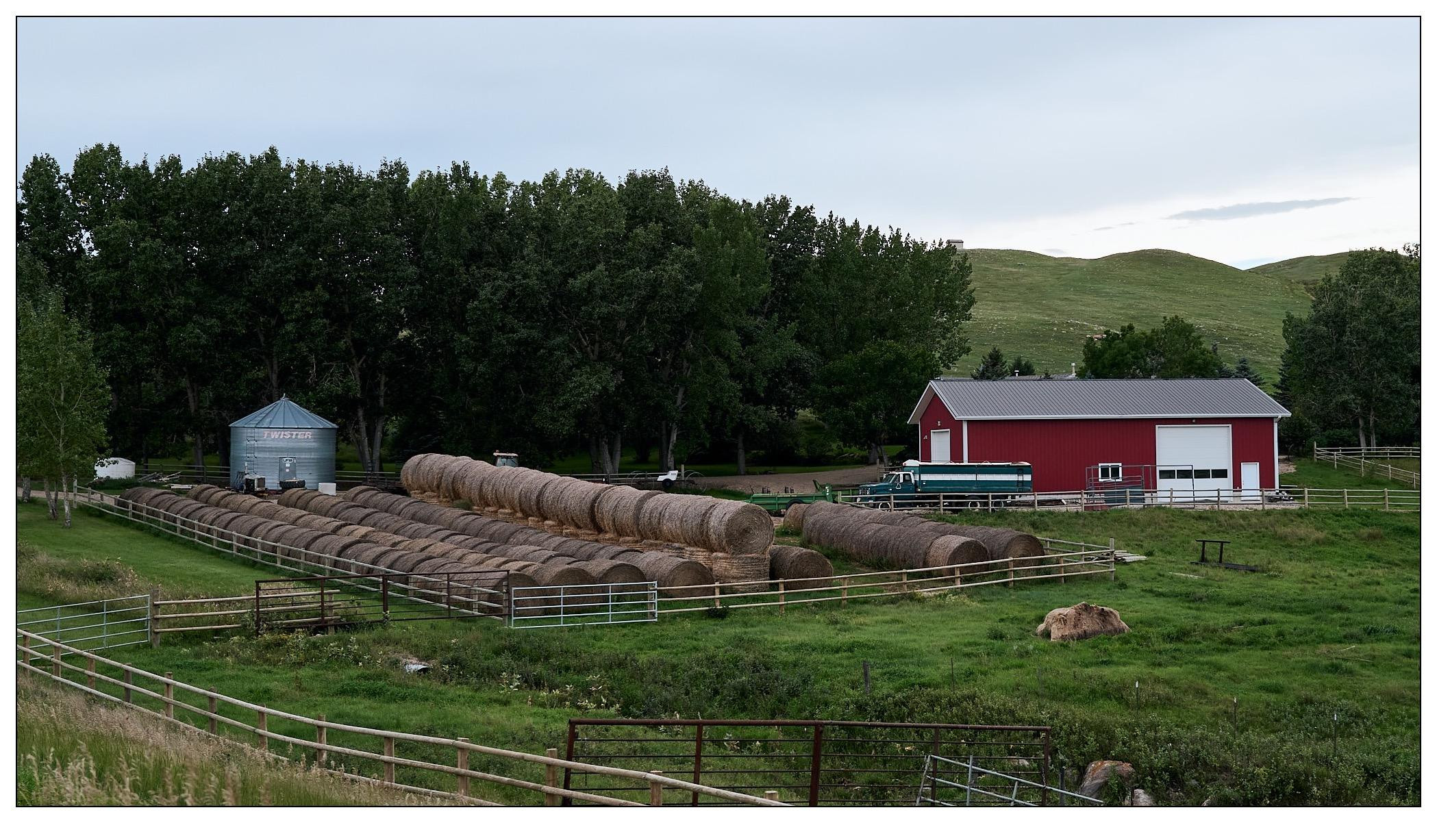 Rural landscape featuring a red barn next to a white steel grain silo and a lineup of large round hay bales. The farm is surrounded by lush green trees with a gently rolling hill in the background. A white fence encloses the area with scattered agricultural machinery visible near the barn.