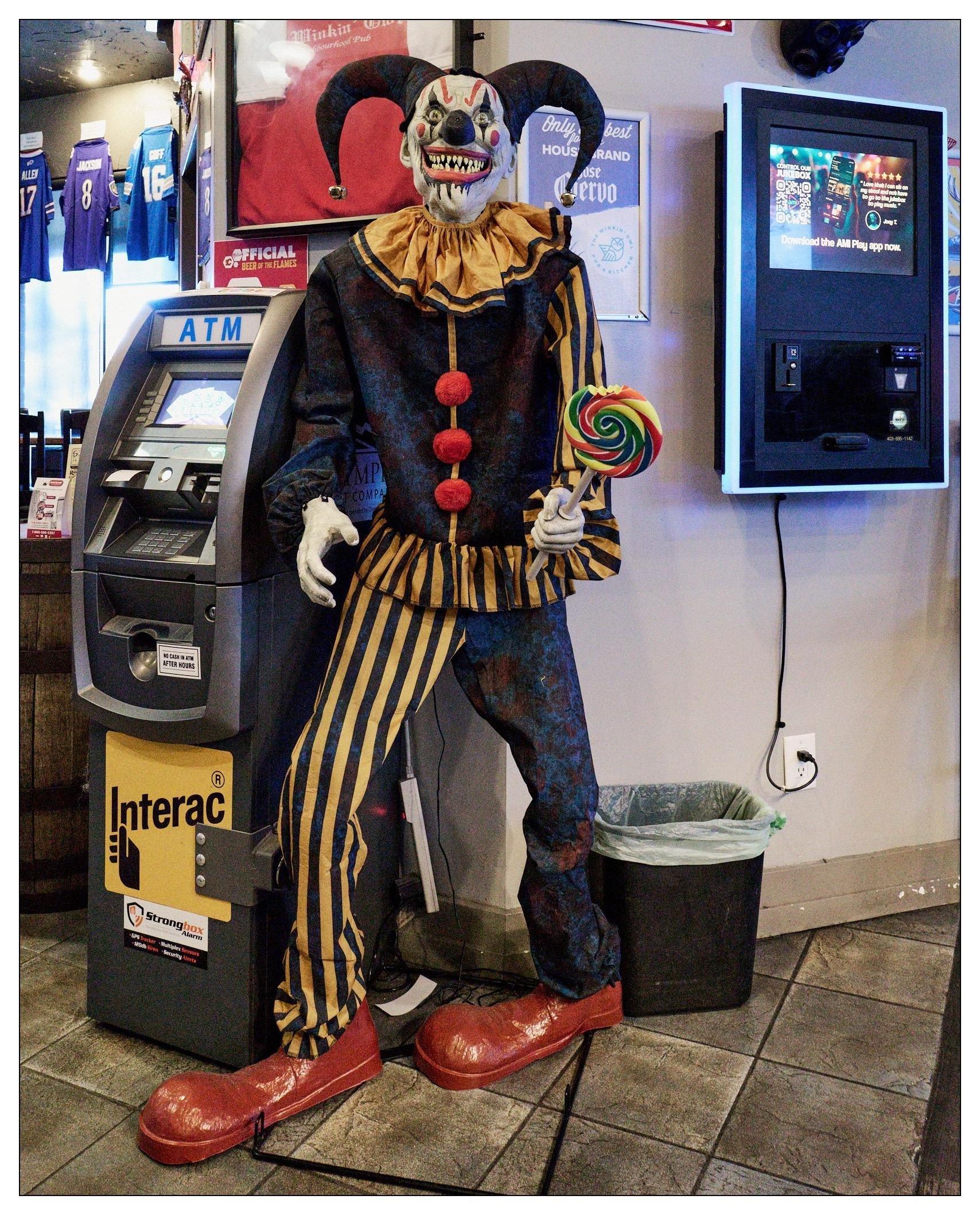 Life-sized clown mannequin dressed in a striped and ruffled clown costume holds a large lollipop in one gloved hand. The clown features an exaggerated, sinister smile with sharp teeth and dark eye makeup, set against large, round ears. It stands in front of an ATM and next to a digital jukebox, under sports jerseys displayed on a wall. A simple black trash can stands next to the mannequin's oversized red shoes on a tiled floor.