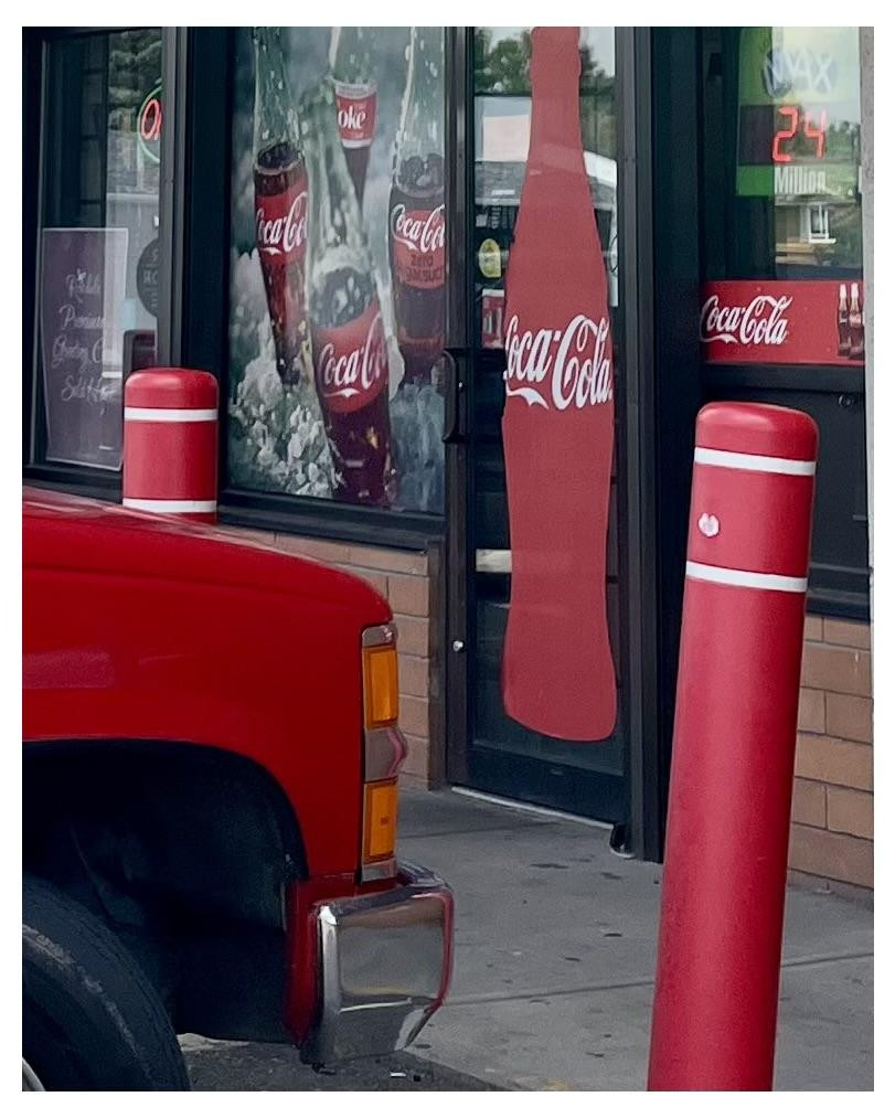 Red pickup truck parked next to two red bollards outside a store with multiple Coca-Cola advertisements visible, including a door shaped like a Coca-Cola bottle and window graphics displaying the soda and ice cubes.