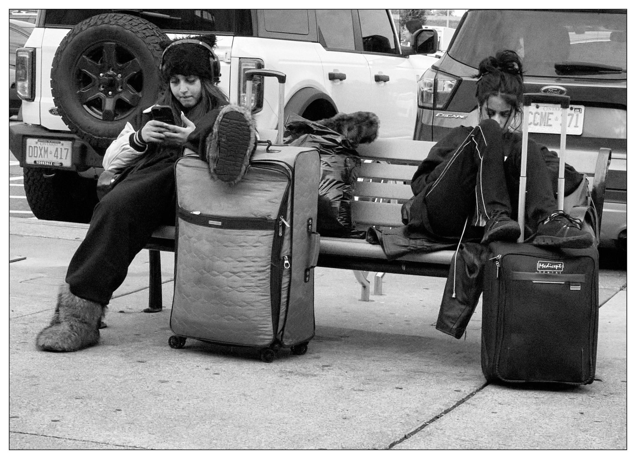 Two individuals, one wearing a fur hat and boots, sitting on a bench with several large suitcases, are engrossed in smartphones in an urban setting with parked vehicles in the background.