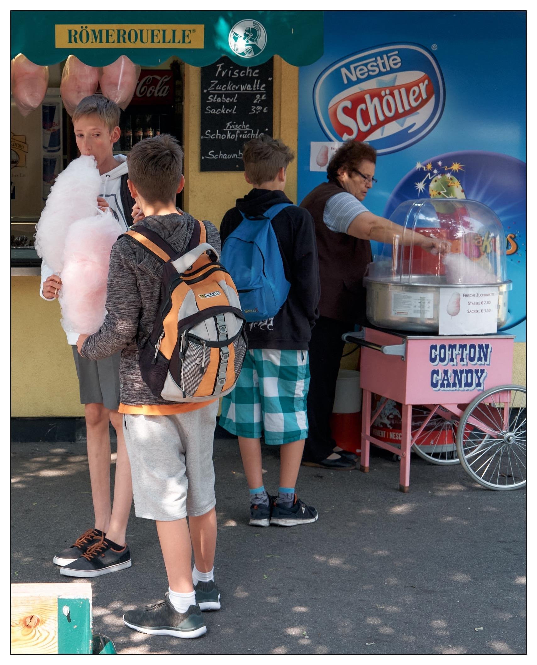 In the image, you see a group of three boys at a cotton candy stall. The boys are holding a large cotton candy each.  The background features signage that includes the brands “Coca-Cola” and “Nestle Schöller”. It appears to be a sunny day and the setting seems to be casual, perhaps at a fair or outdoor market. The atmosphere is lively and colorful, suggestive of a leisurely, enjoyable day out.