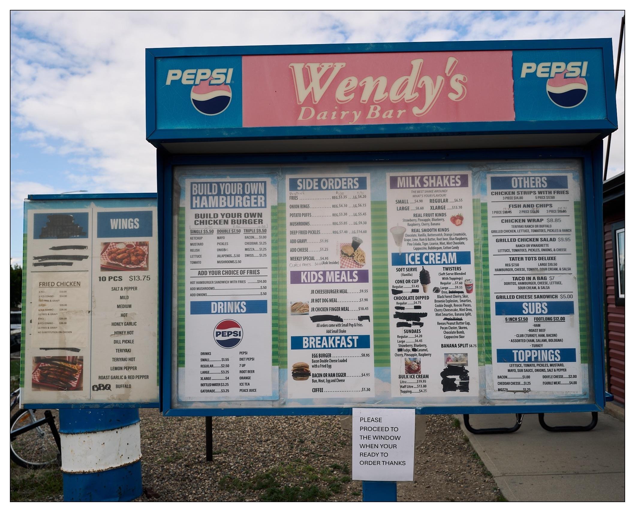 Close up of the menu sign, which is labelled "Wendy's Dairy Bar".  It has many items blacked out and an auxiliary menu added to the side with other things like chicken wings.