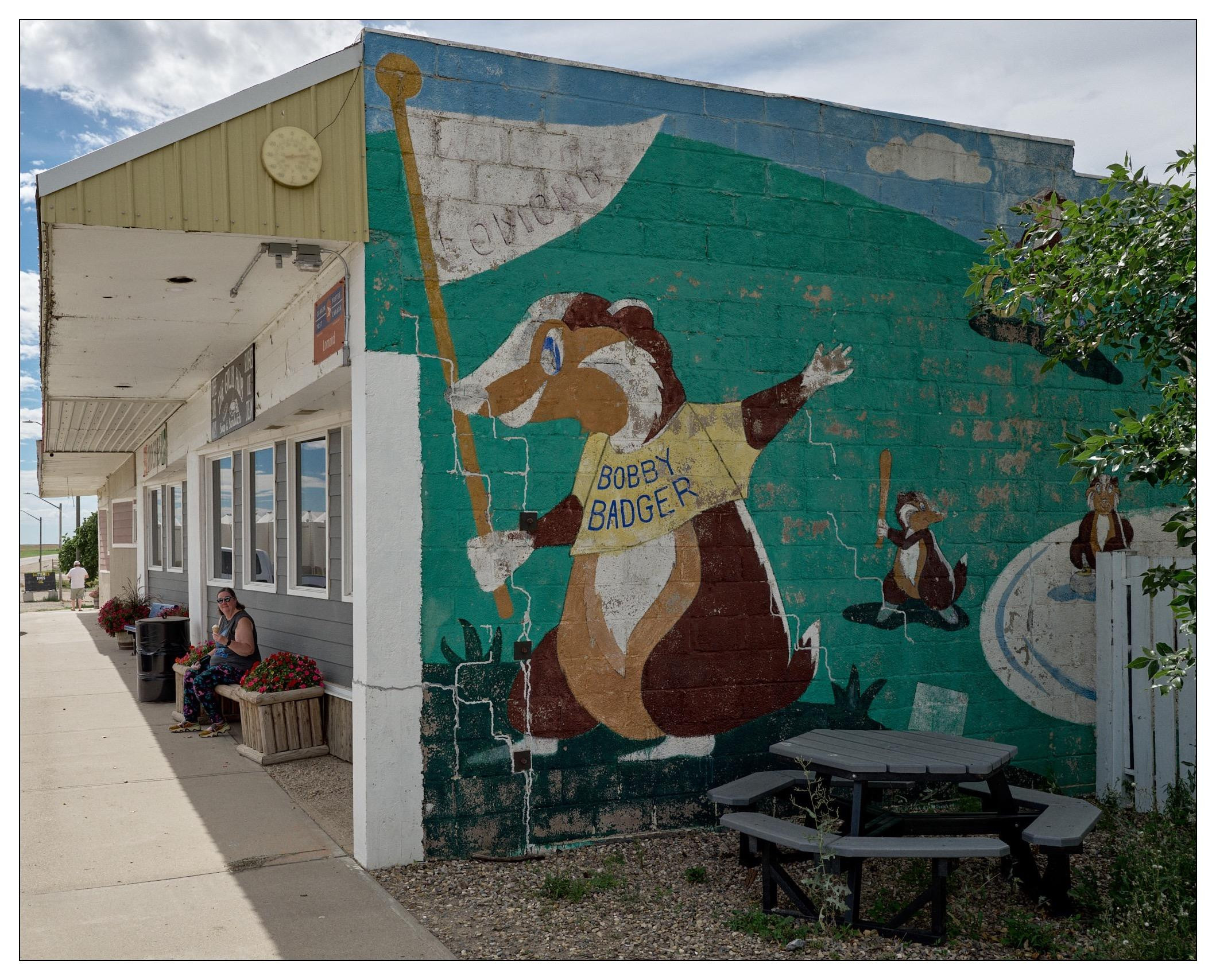 Mural of a cartoon badger dressed in a yellow shirt labeled "Bobby Badger," waving a white flag, on a green wall beside a building with an extended roof sheltering benches and a seated person. The scene includes a small wooden picnic table in front, and a sparse tree slightly obscuring the mural. The mural is weathered, showing cracks and paint peeling, suggesting it is an older artwork.