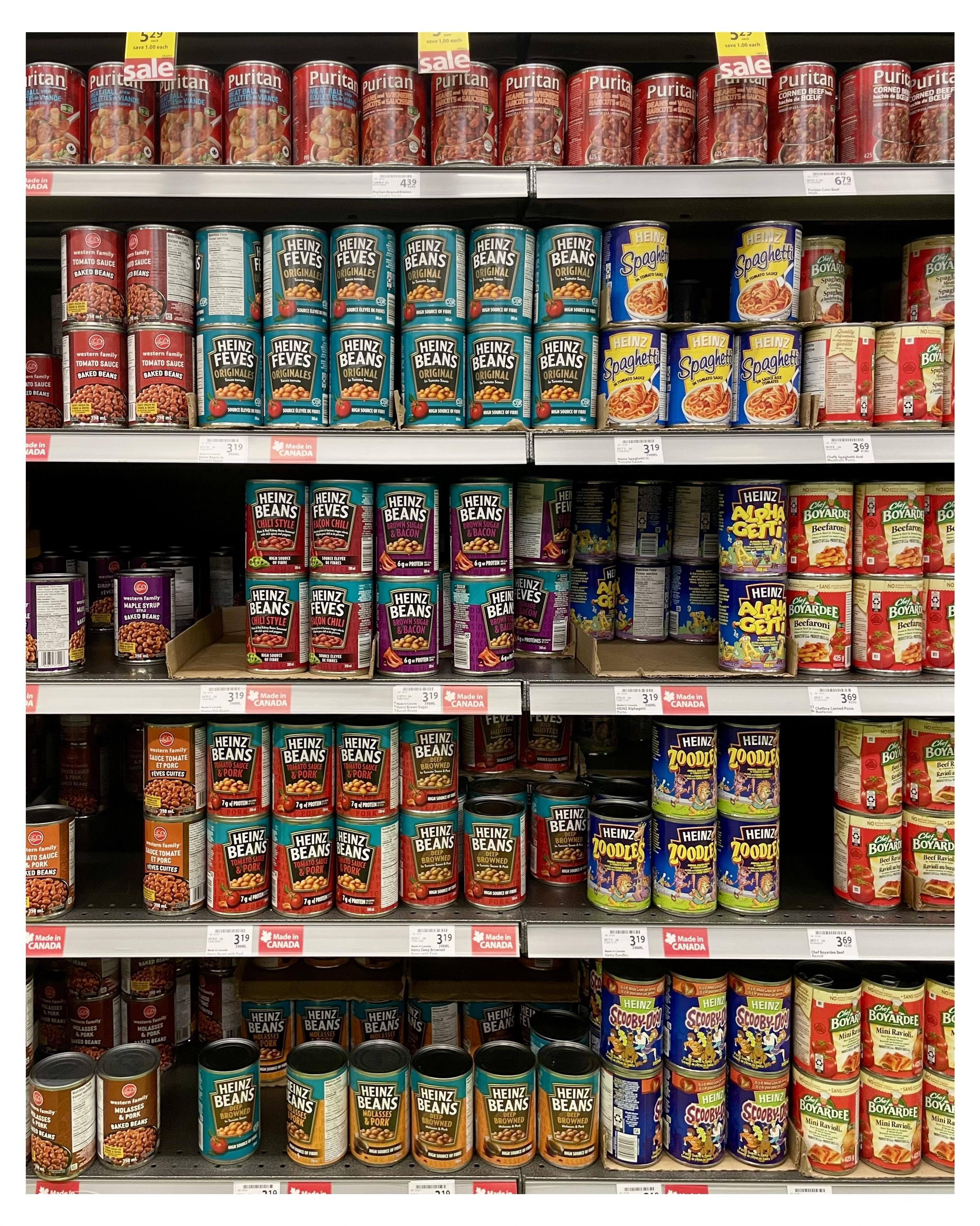 Canned food products, predominantly beans and spaghetti variations, are organized on supermarket shelves. Brands include Heinz and Puritan prominently displayed with labels facing forward. The shelves are stocked in tight rows, each can positioned next to similar products, with some marked on sale. Prices and product names are visible on small shelf tags below each group.