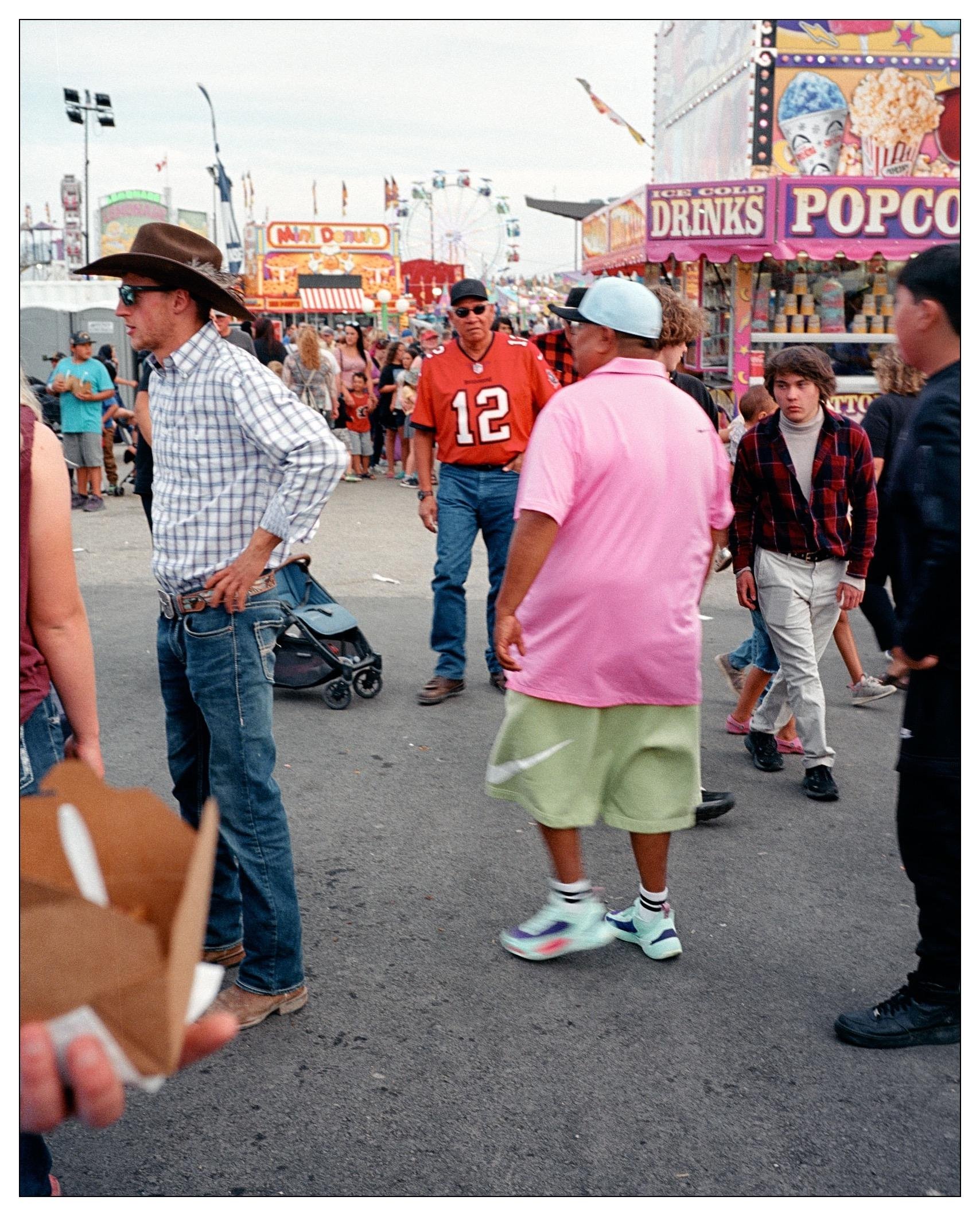 This image captures a lively scene at a fair or carnival. You can see a variety of people dressed in different styles, suggesting a diverse crowd, and the environment is bustling with activity. Notable features include:

1. A man wearing a cowboy hat, sunglasses, and a plaid shirt on the left side mid-turn, seemingly gazing at something outside the frame.
2. A person in a bright pink shirt and green shorts, facing away and towards the center of the image.
3. A man wearing sports jerseys (one in an orange jersey with the number 12), which suggest he might be a fan of a sports team.
4. A variety of carnival booths in the background advertising popcorn and ice-cold drinks.
5. Many other fairgoers walking around or engaged in conversation.

Overall, the image depicts the vibrant atmosphere of a public event with various individuals enjoying their day out.