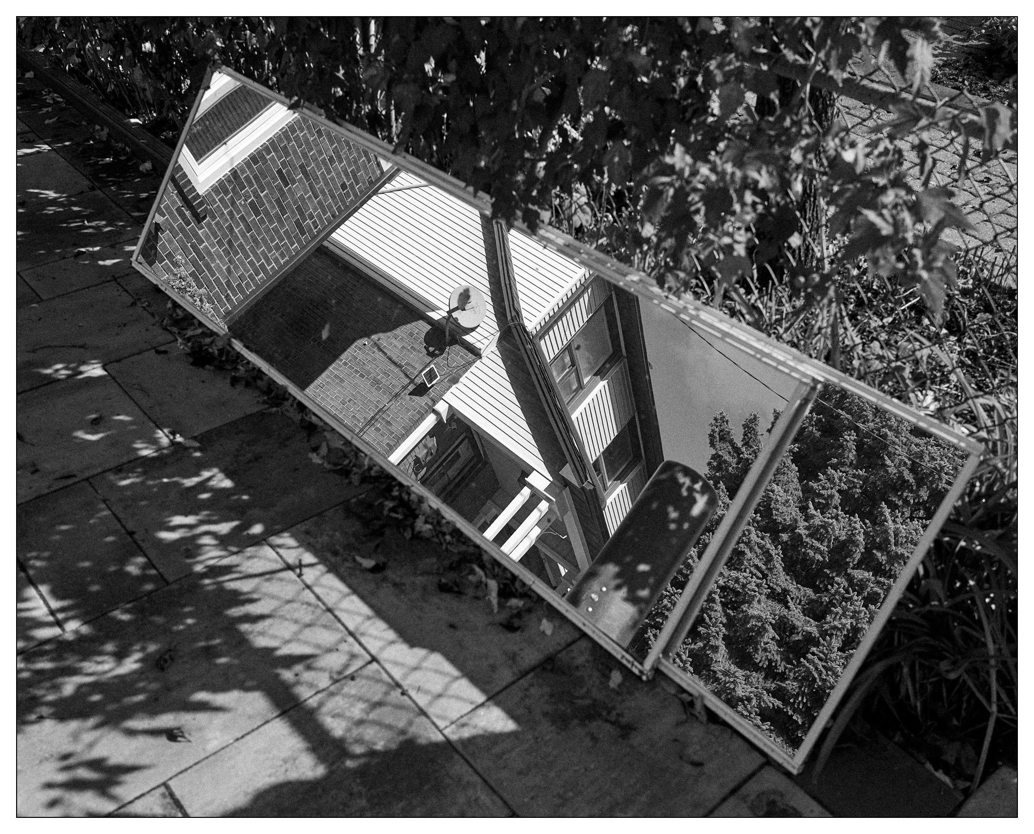 Black and white photograph of a passerby's reflection in a tilted, rectangular mirror lying on a sidewalk. The mirror reflects an adjacent building with a door, satellite dish, and a porch, surrounded by trees and partly obscured by overhanging leaves. Shadows of leaves pattern the ground around the mirror.