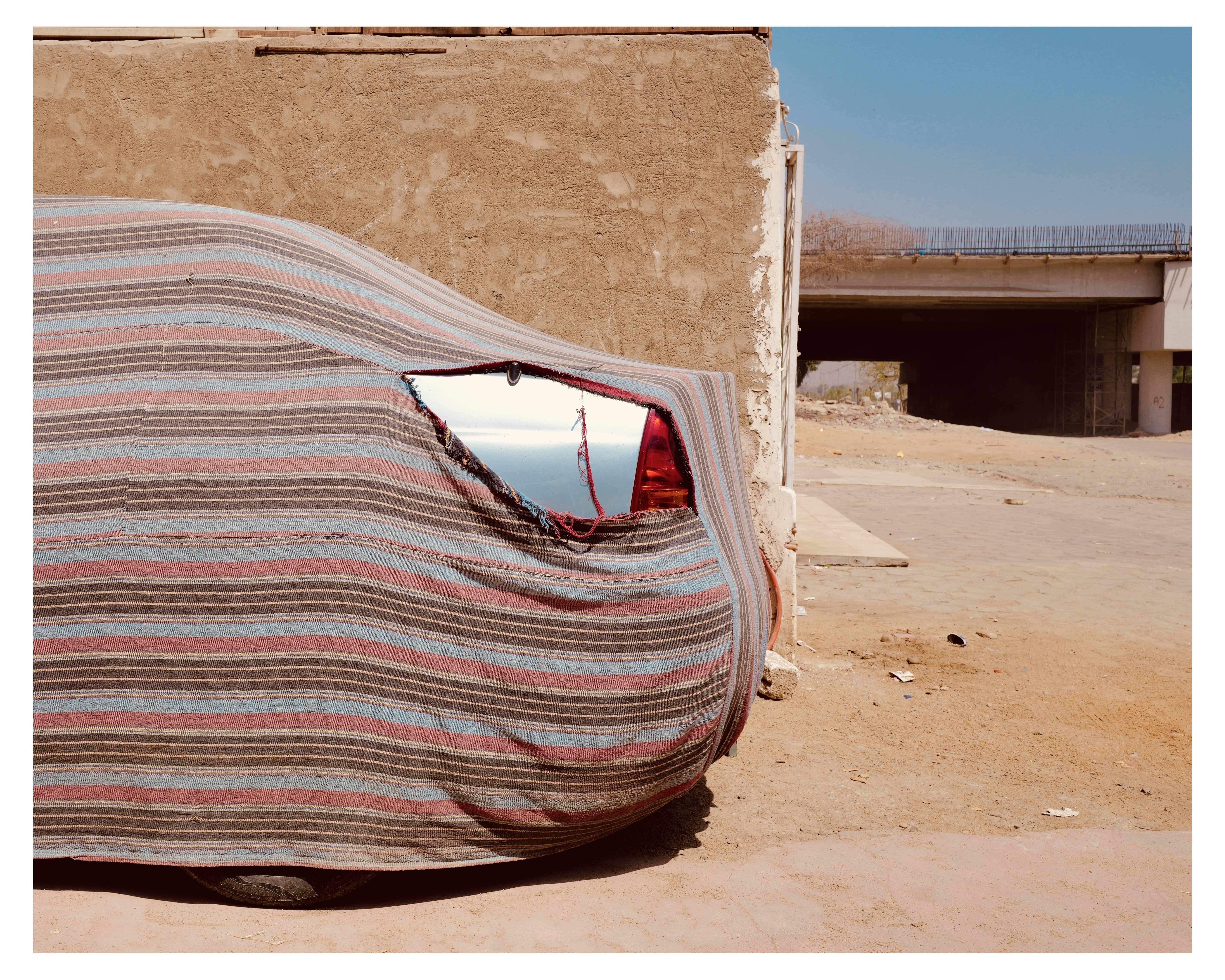 The image shows a car covered with a striped fabric cover, parked on a dirt space next to a beige-colored building wall. The cover is slightly torn at the back, revealing the rear of the car which includes a shiny, possibly red painted surface and a taillight. In the background, there is a bridge or an overpass structure. The scene appears to be in a dry, possibly urban environment given the concrete structures and absence of vegetation.