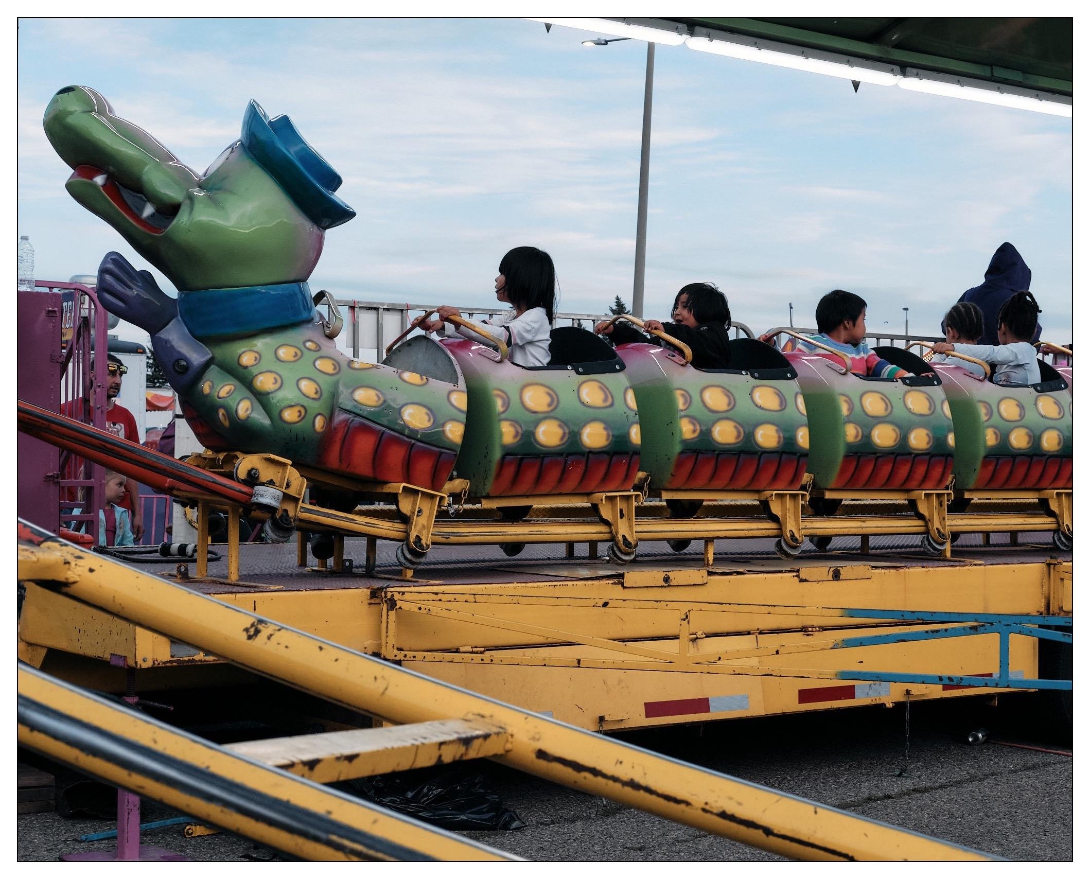 This image captures a colorful amusement park ride resembling an alligator. The structure includes a segmented body in yellow, green, and red with circular patterns, a smiling face, and a hat-like element on the head adding a whimsical touch. Children are seated in the compartments of the ride, secured by safety bars, and are seemingly enjoying their time as the ride operates on a track. The scene is set outdoors under an overcast sky, contributing to the mild lighting in the image. In the background, a supervising adult and some operational details of the ride, like stairs and control panels, are visible.