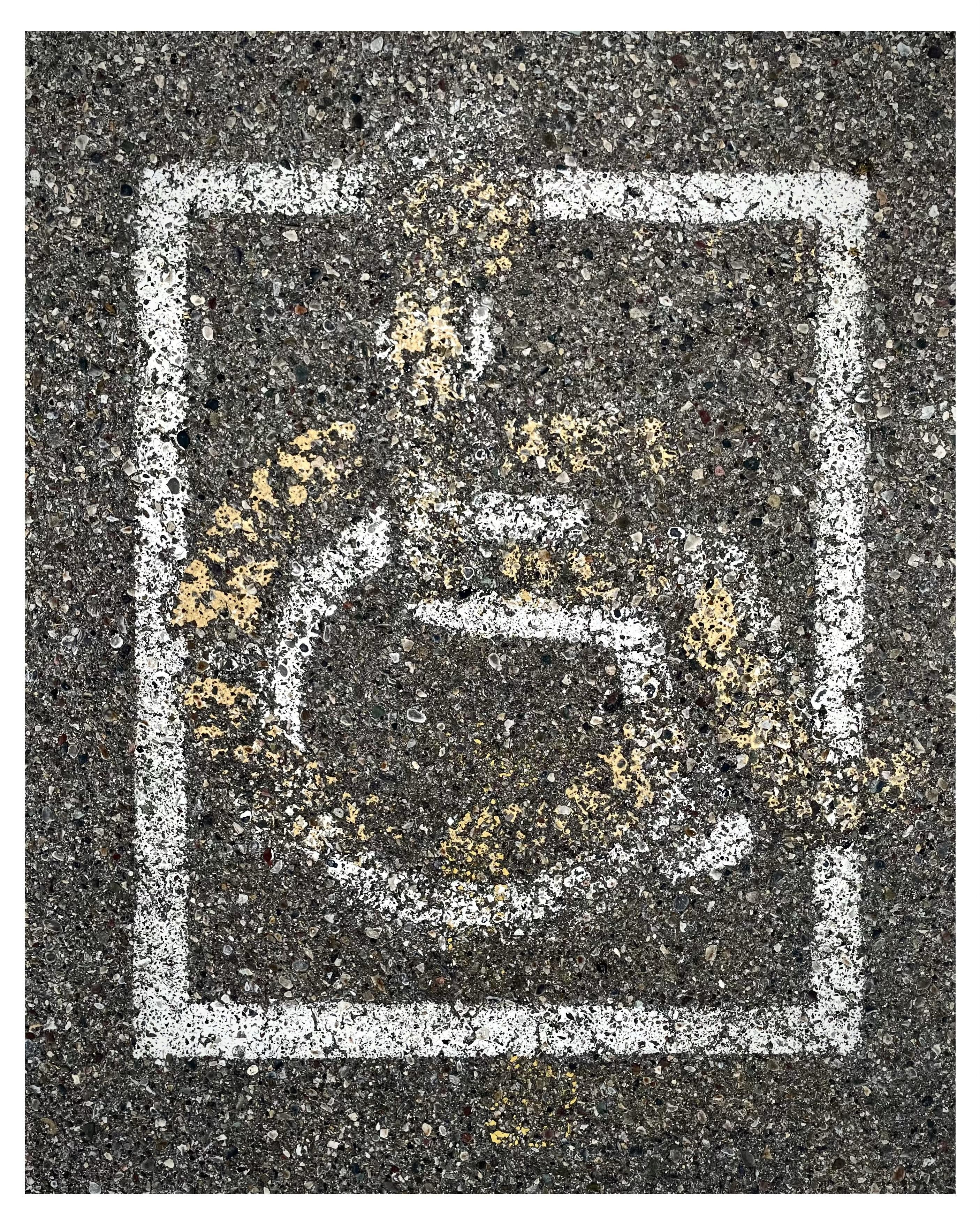 Faded white painted disabled parking stencil enclosed in a square on a textured asphalt surface, showing signs of wear and partially covered by a faded yellow stencil of slightly different design.