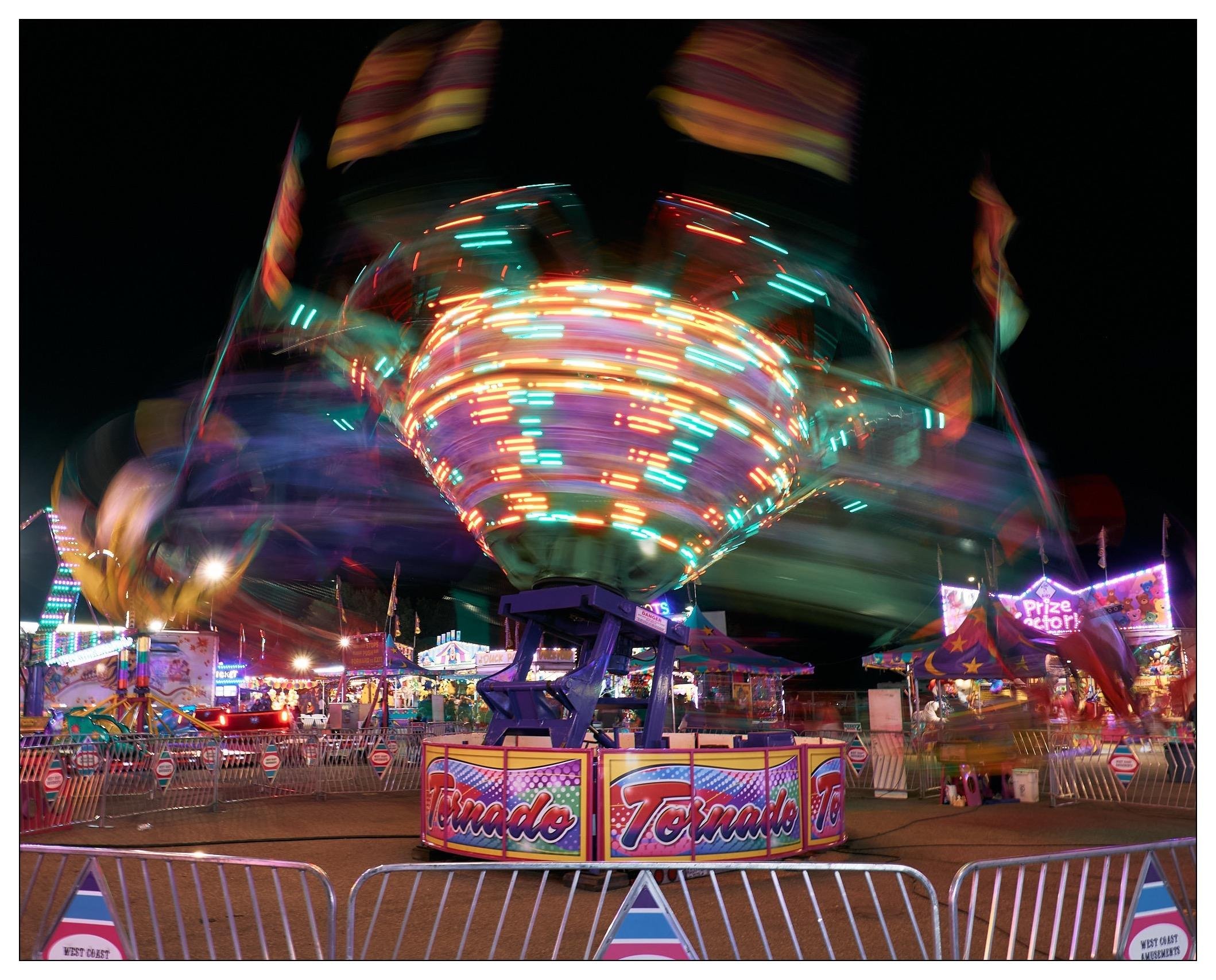 This image captures a vivid scene from a carnival or fair at night. It features a rapidly spinning ride called "Tornado," which is brightly illuminated with colorful lights creating a dynamic, blurry motion effect. The background shows various other carnival attractions, games, and booths, also adorned with lights, contributing to the festive atmosphere. Metal barriers are in the foreground to ensure the safety of spectators and potential riders. The overall image conveys the excitement and energy typical of a busy fairground.
