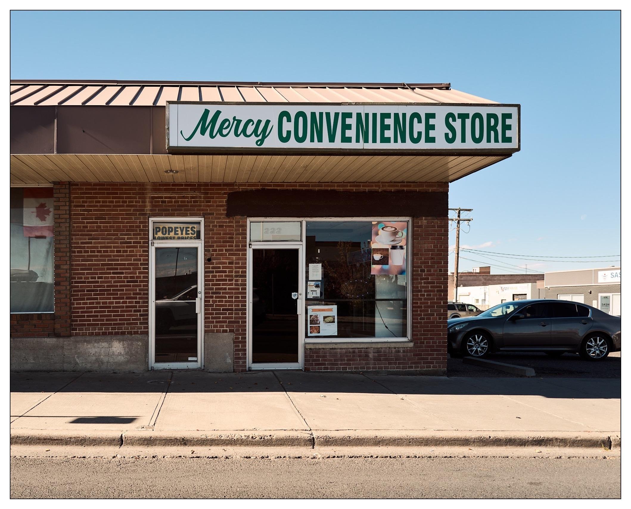Exterior of Mercy Convenience Store with a green and white sign above the entrance. The single-story brick building has large glass windows displaying advertisements and a transparent door. A sign for Popeyes is also visible on the window beside the door. The store is set against a clear sky, with a parking area visible on the right where cars are parked.