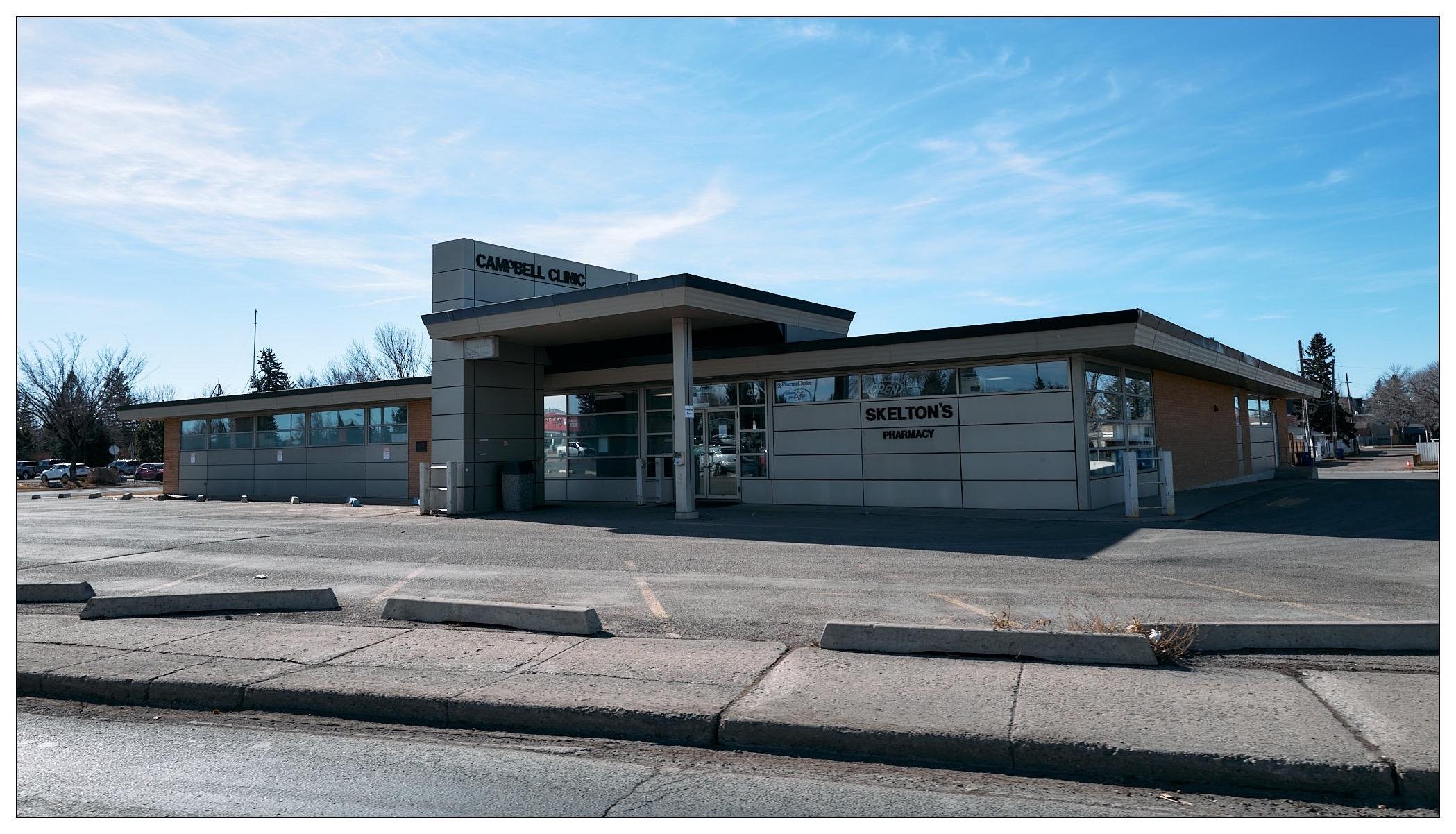 A modern, single-story commercial building that houses a medical clinic named "Campbell Clinic" and a pharmacy named "Skelton's Pharmacy." The building features a flat roof, large glass windows, and a minimalistic architecture style. The setting appears to be an urban or suburban area with a parking lot in the foreground, and it seems to be a clear sunny day with few clouds in the sky.