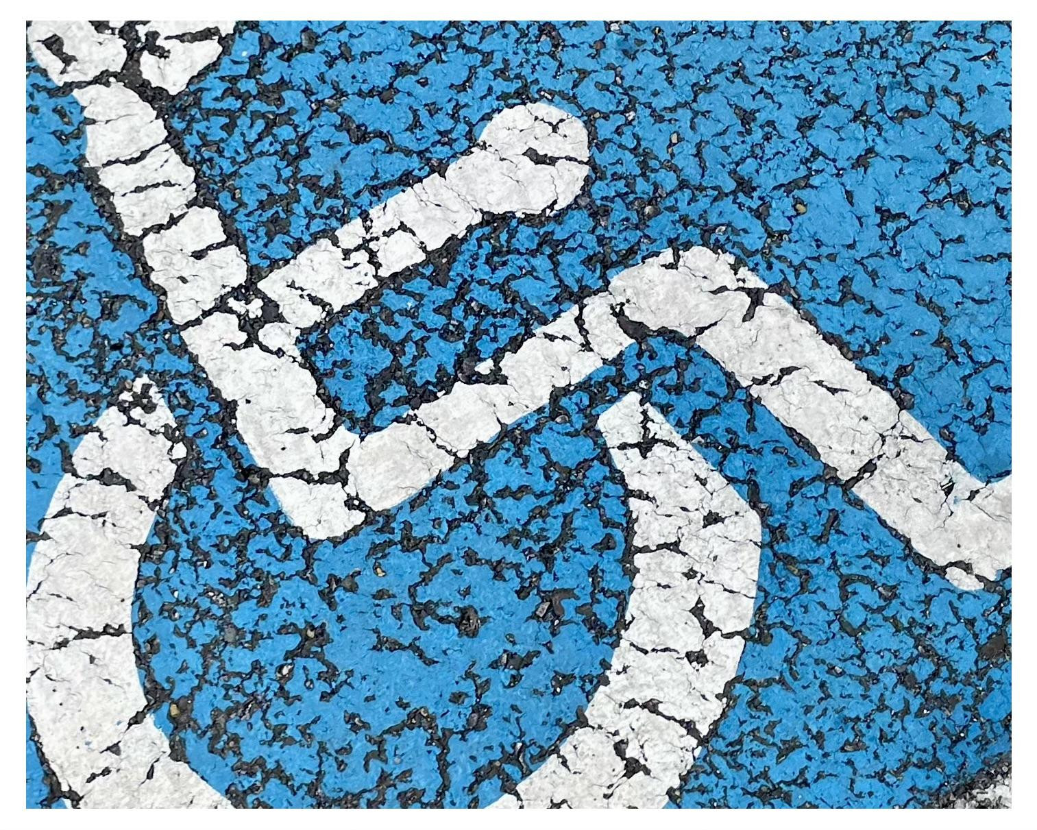 Close-up view of a weathered blue painted surface with a white wheelchair symbol, showing cracked paint and the underlying asphalt texture.