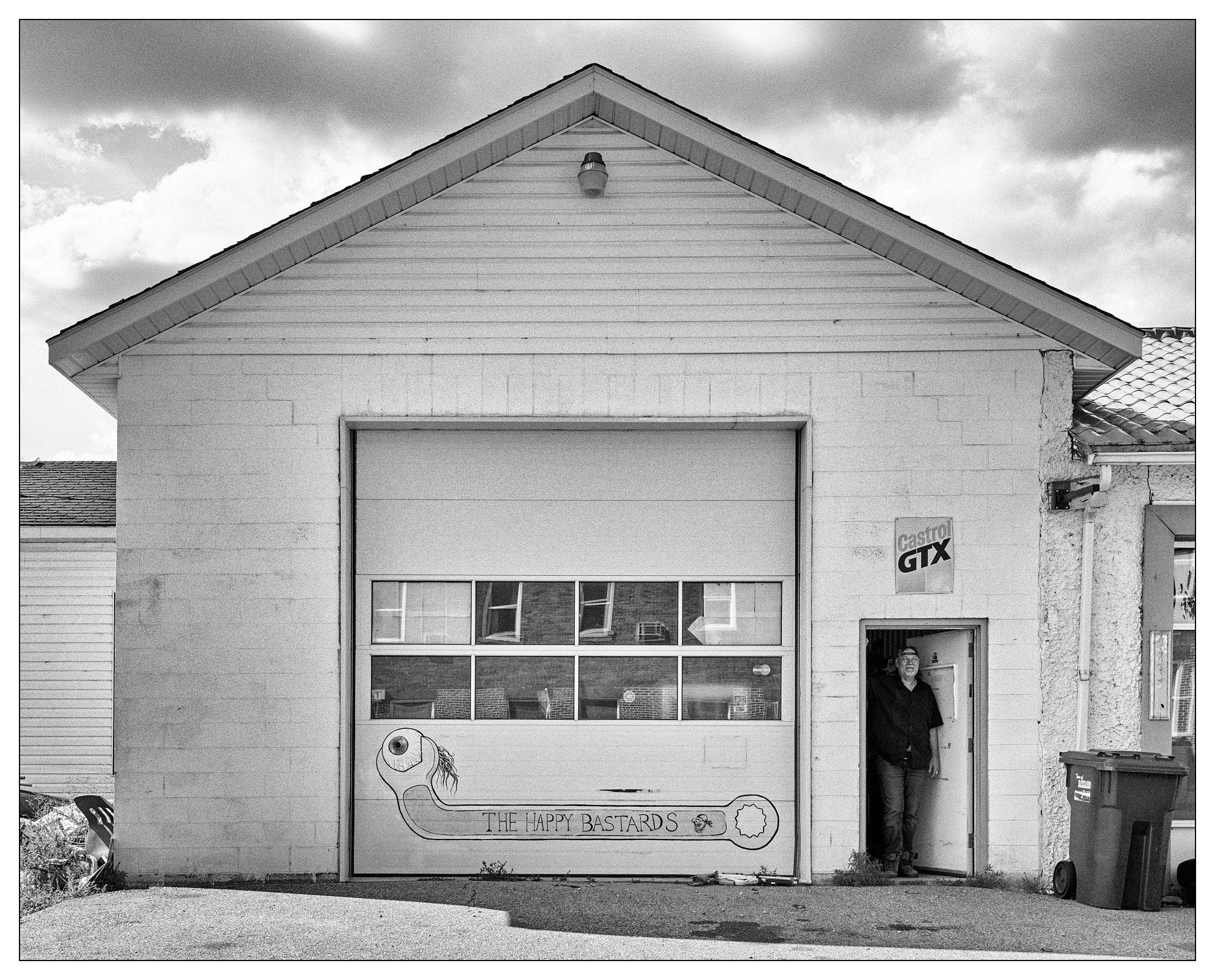 Black and white photo of a rectangular white garage with a large door featuring a whimsical drawing of a fish and the words "The Happy Bastards." Reflective windows and a small lantern adorn the gabled roof. A man stands in the doorway on the right side of the garage, next to a sign advertising "Castrol GTX." A trash bin is also visible beside the man.