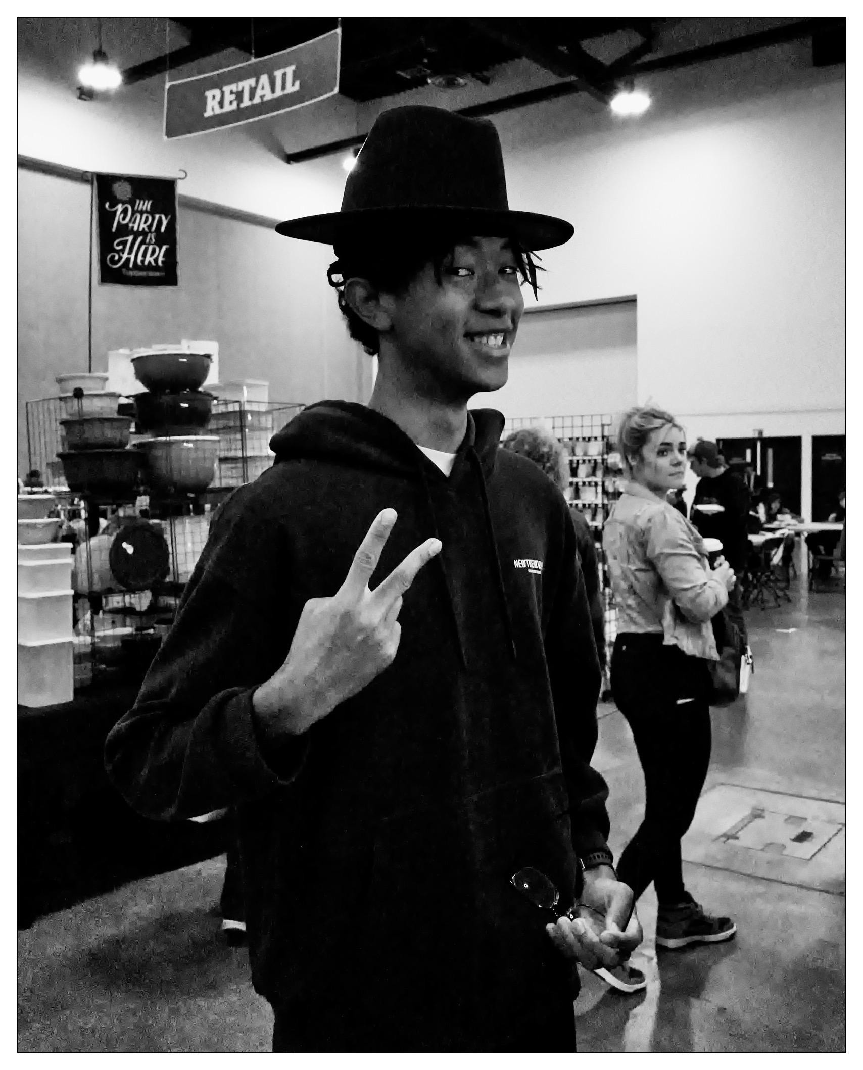 Black and white photo of a smiling young man wearing a dark wide-brimmed hat and a black hooded sweatshirt labeled "VANS". He gestures a peace sign with his right hand. In the background, there are banners labeled "RETAIL" and "THE PARTY IS HERE" in an indoor setting, possibly a convention or market, with other attendees visible.