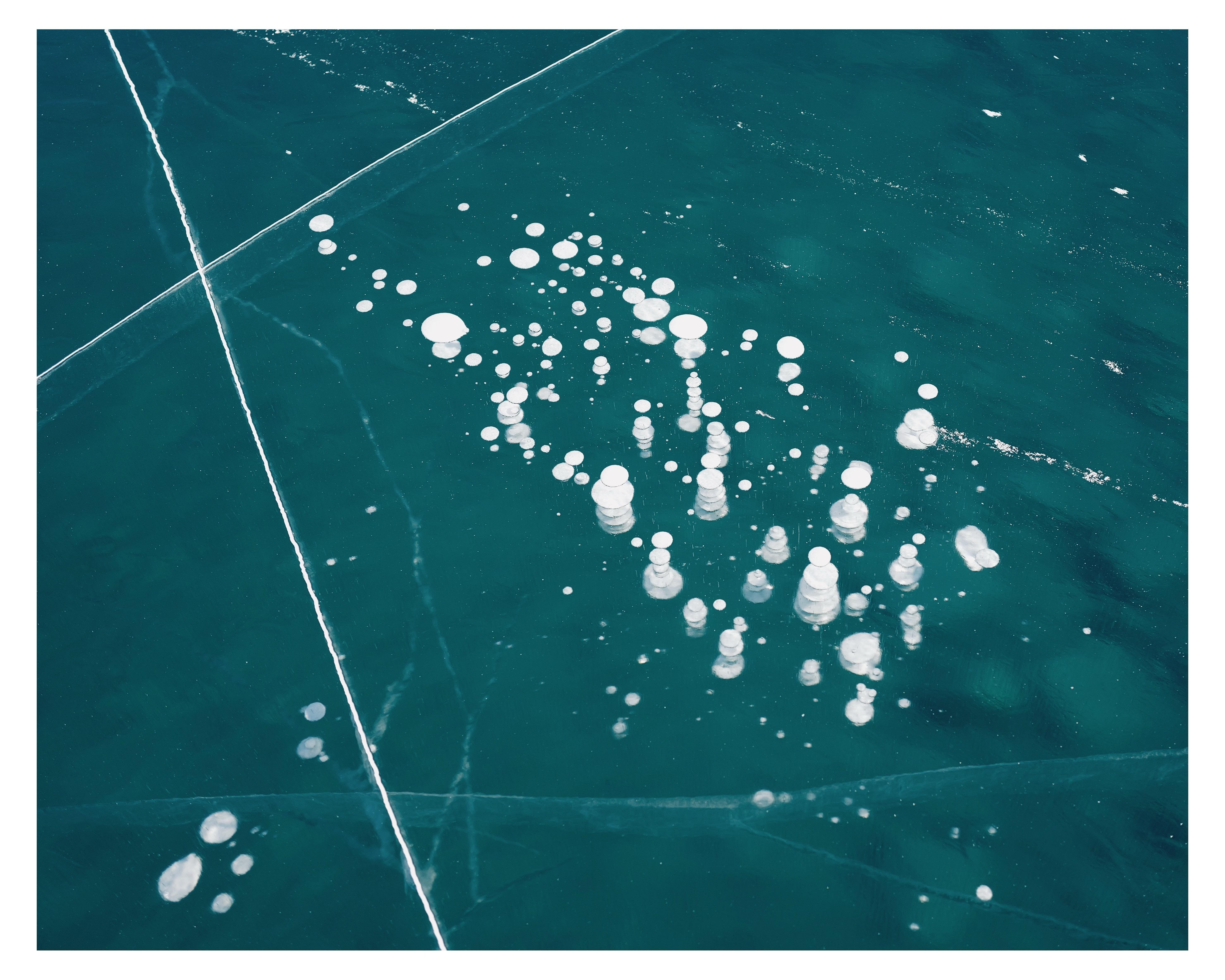 Frozen water surface with a scattering of variously sized white bubble formations trapped under the clear ice, with visible cracks intersecting the bubbles.