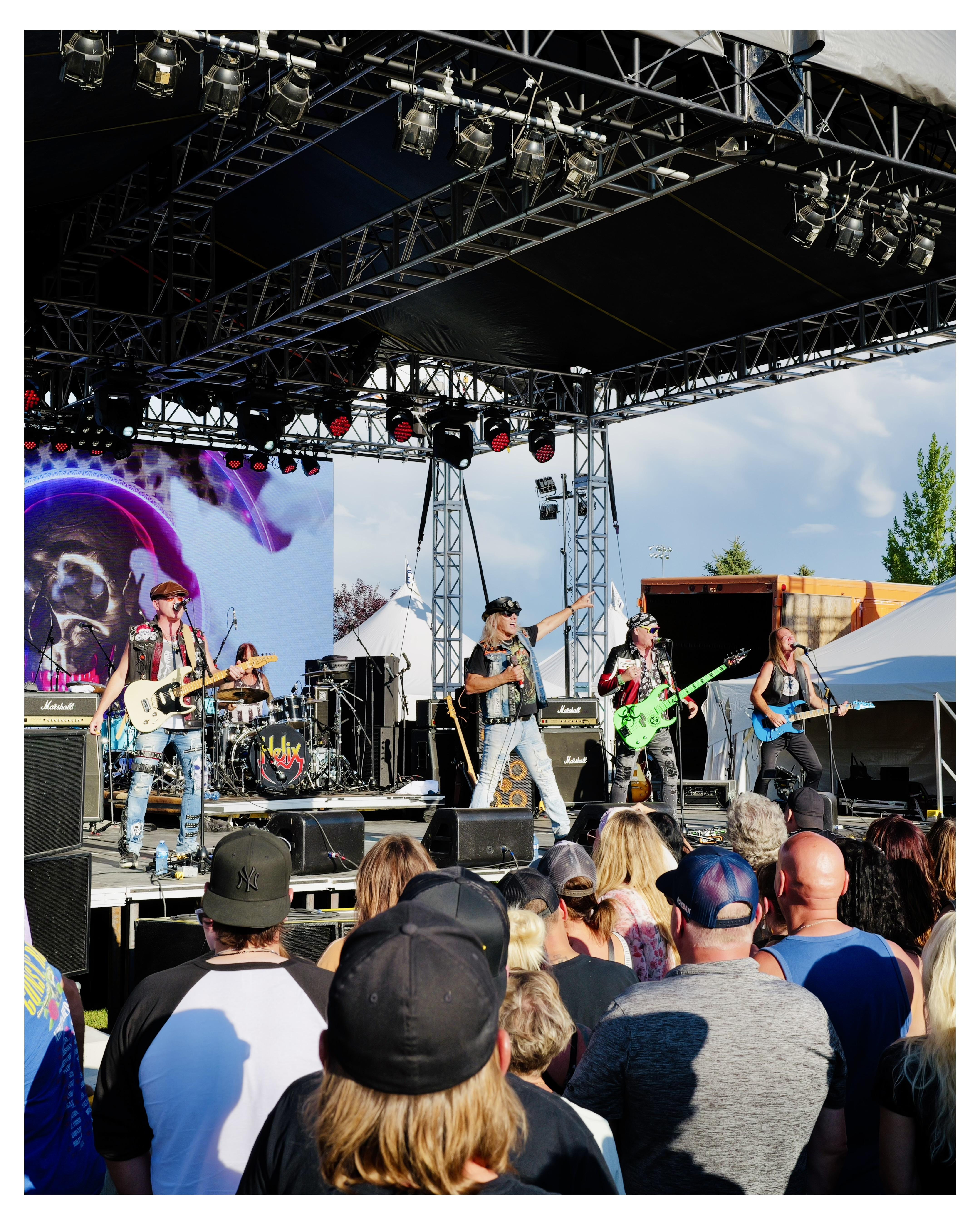 Outdoor concert with a band performing on stage, with four musicians: two guitarists, a bass player, and a drummer. The stage features an overhead lighting rig and a large screen displaying a purple-tinted image of a skull wearing sunglasses. Audience members, viewed from behind, are standing and watching the performance, some wearing hats and casual clothing.