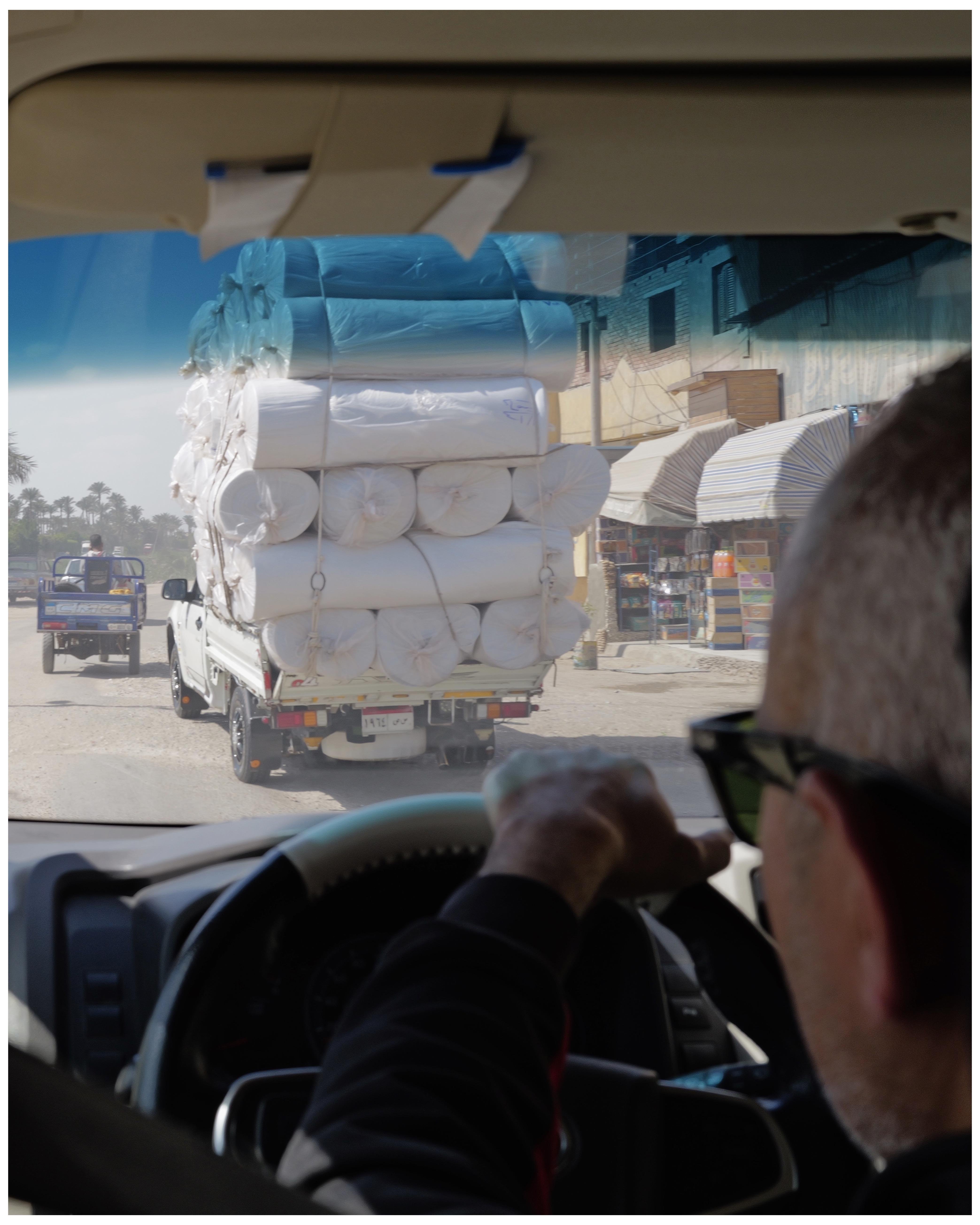 This image shows a view from inside a vehicle, capturing another vehicle in front which is heavily loaded with various rolls, possibly of fabric or some other material. The cargo is tightly wrapped, all secured with straps. Viewed through the windshield, part of the driver, who appears to be wearing sunglasses and gripping the steering wheel, is also visible. Further ahead, other vehicles and roadside shops can be seen, suggesting an urban setting.