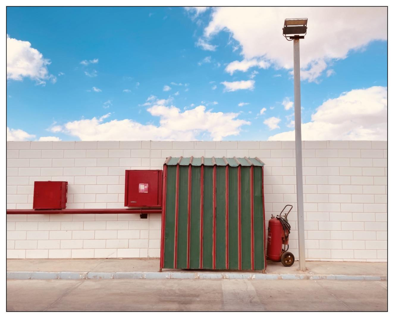 The image shows an outdoor wall that appears to be part of an industrial or utilitarian building. The wall is composed of light colored bricks. Attached to the wall are three red items: two square-shaped boxes (perhaps electrical boxes or alarm systems) and a corrugated metal door painted in alternating vertical stripes of green and red. On the ground next to the wall sits a portable red fire extinguisher with wheels. A tall light pole stands near the right edge of the image. The sky above is blue with some scattered clouds, suggesting a clear weather day.