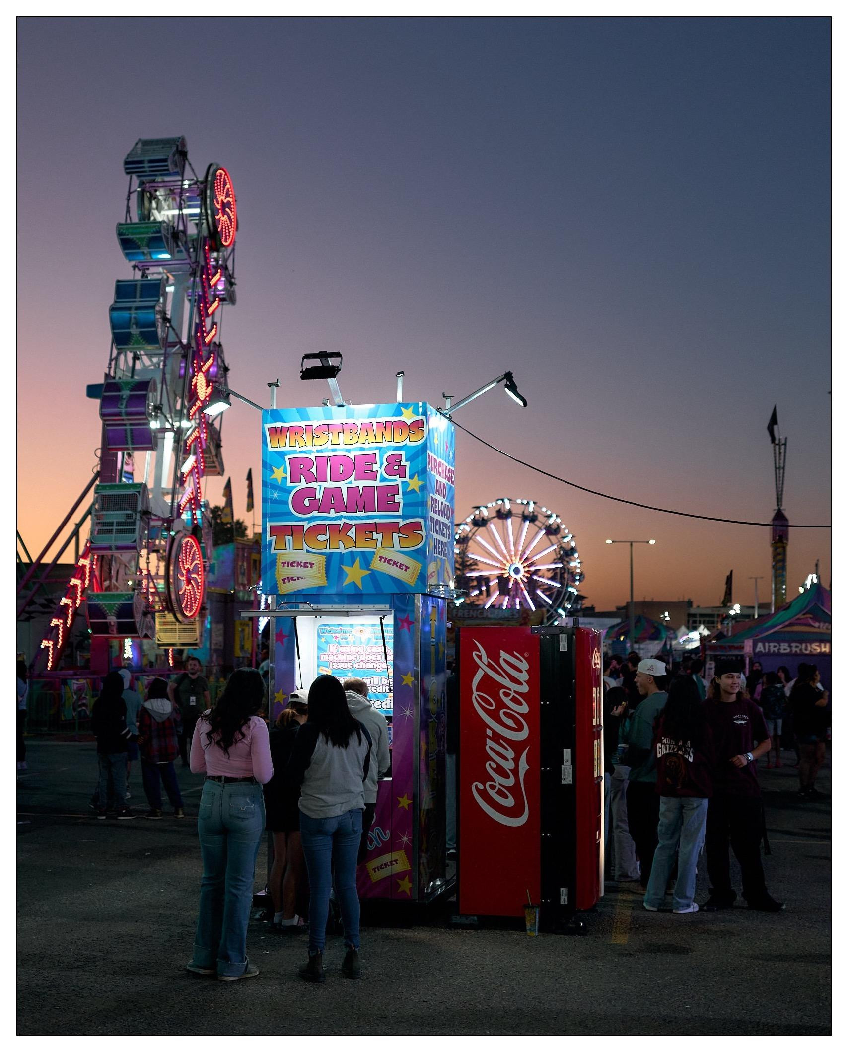 Evening view of a carnival with a vibrant atmosphere, featuring people gathered around ticket booths labeled "WRISTBANDS RIDE & GAME TICKETS". Prominent in the background are illuminated amusement rides, including a Ferris wheel and a vertical rotating ride, set against a twilight sky. To the right, individuals are engaged in conversation near a Coca-Cola vending machine. The scene captures the glow of neon lights and the festive environment typical of a carnival setting.
