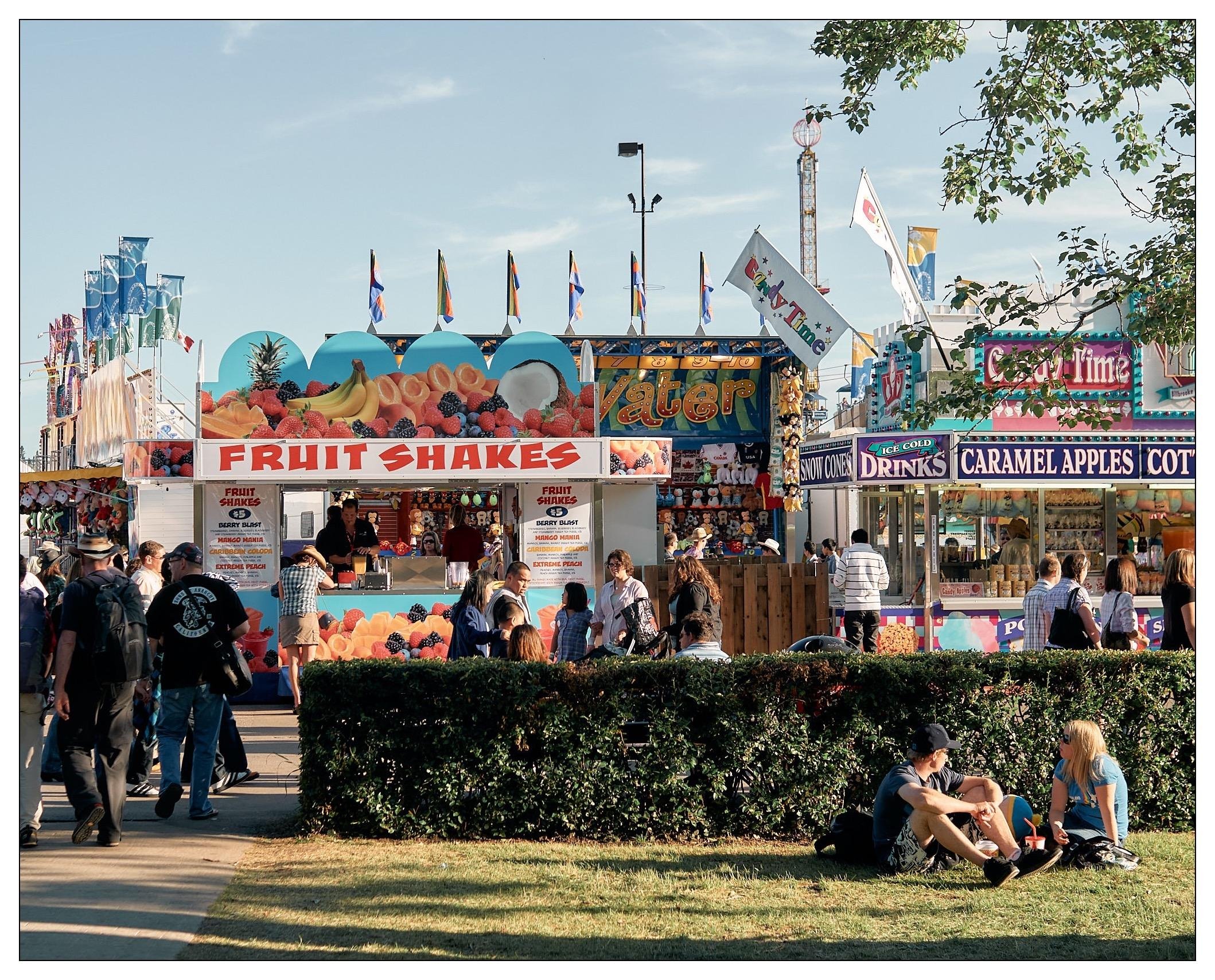 The image shows a bustling fairground with various food and drink stalls. A prominent stall titled "Fruit Shakes" is featured in the foreground, selling fresh fruit shakes, as suggested by images of colorful fruits displayed on its facade. Other stalls visible in the background offer a variety of treats, including "Drinks," "Caramel Apples," "Cotton Candy," and more, highlighting the typical fairground snacks.

The scene is lively, with many people walking around, visiting the stalls, or sitting and relaxing on the grass. There's also a decorative hedge in the foreground, contributing to the festive and orderly environment. In the distance, you can see amusement rides and flags, enhancing the fair's vibrant atmosphere. The setting indicates a sunny day, perfect for outdoor activities like a fair.