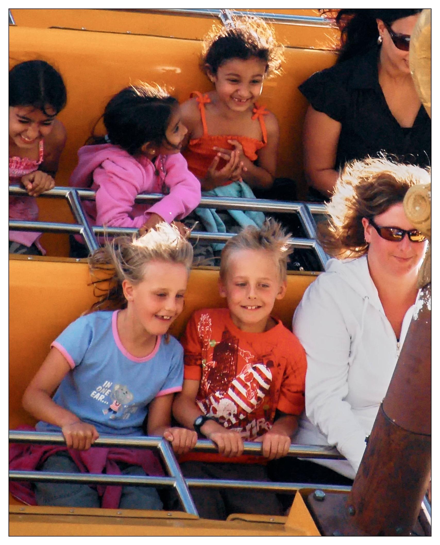 This image shows a group of children and adults enjoying a ride, on a rocking ship. The children look excited and joyful, and some are smiling broadly at the thrill of the ride. They are seated in a brightly colored yellow carriage with metallic safety bars. The setting suggests a fun, lively day out at an amusement park, capturing a moment of pure childlike happiness and adventure.