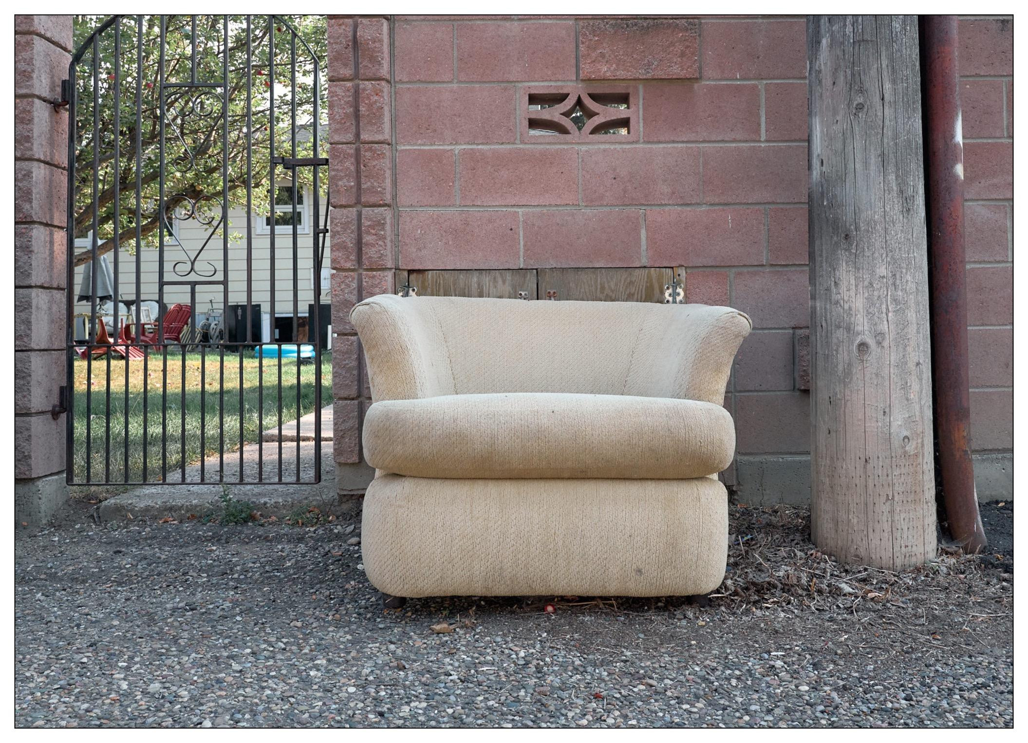 Beige armchair placed against a brick wall next to a metal gate leading to a yard with a visible swing set and pool. A wooden post stands adjacent to the chair on the right, partially framing a house in the background.