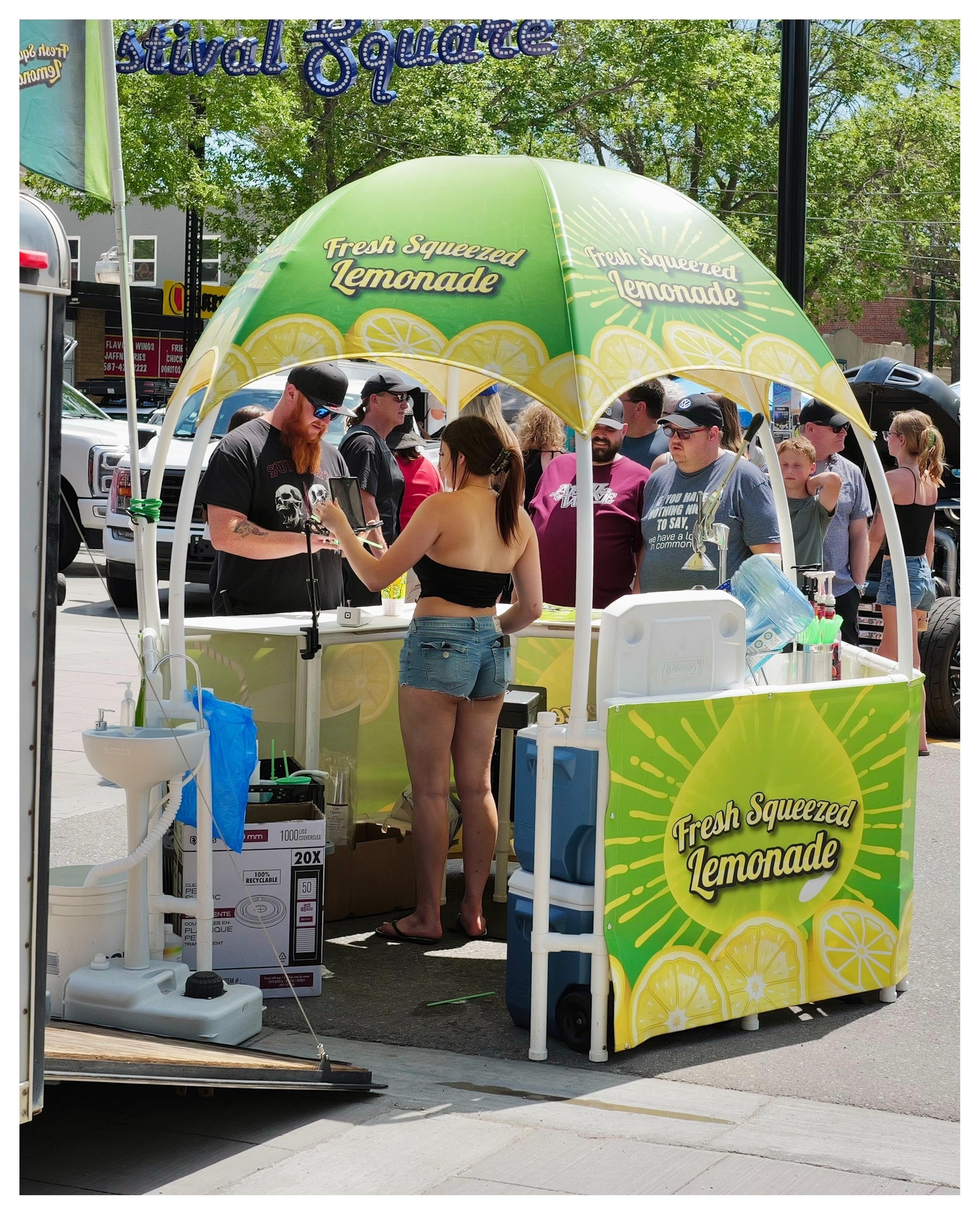 Rear view of lemonade stand at an event, we can see the inside details including the woman running the booth.   She wearing a black baseball cap, black tube top and jeans short shorts.  Her butt cheeks are hanging out.  A crowd of men are on the front side looking in, getting their drinks.