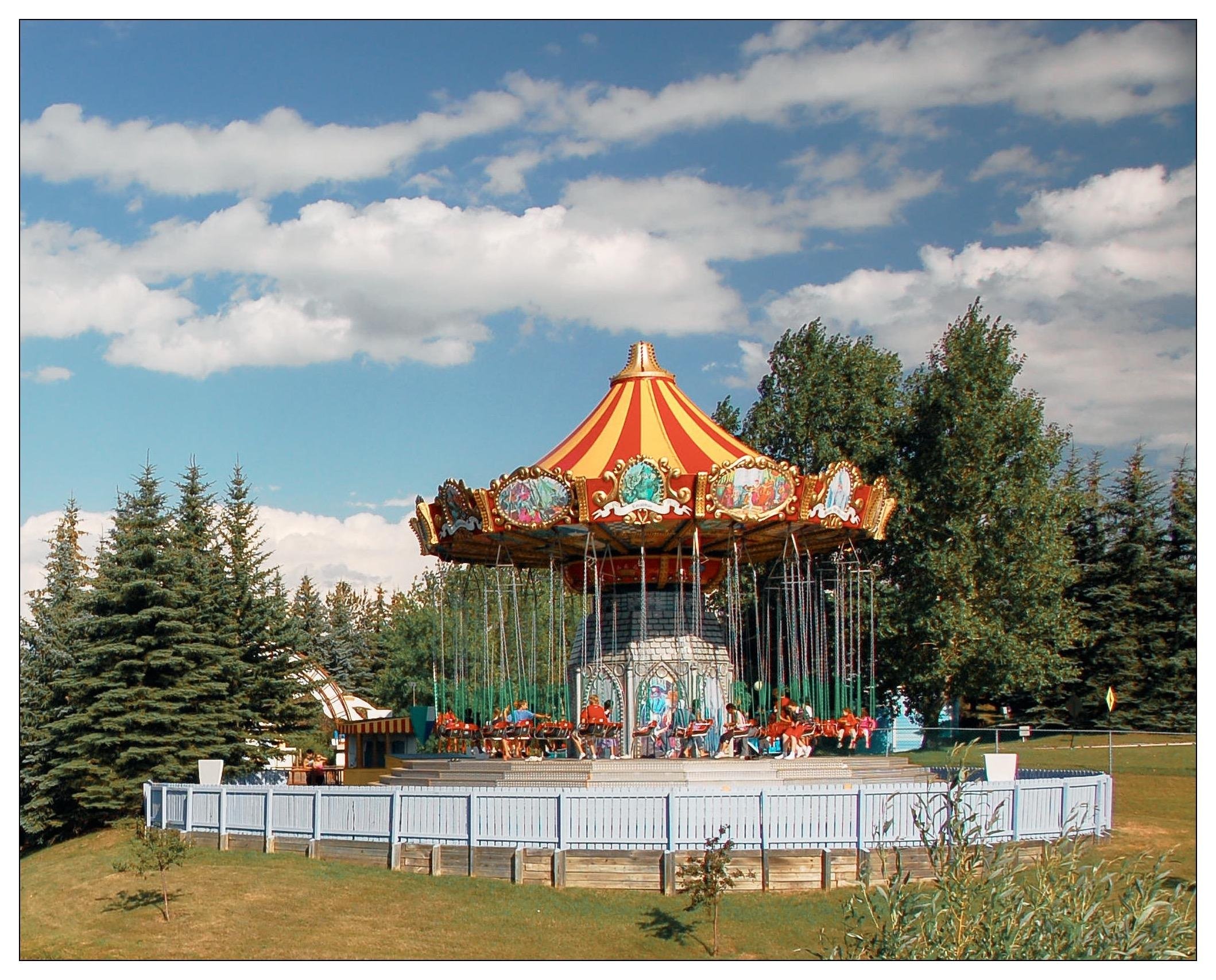 This image shows a traditional outdoor carousel ride at an amusement park. The ride features a large, colorful canopy with decorative elements, and suspended chairs that are designed for riders to sit in while they swing outwards as the carousel spins. The setting includes a blue sky with puffy clouds and a landscape with mature trees that suggests a well-maintained park or recreational area. Surrounding the carousel are protective railings and a structured enclosure, with the overall scene conveying a joyful, leisurely atmosphere typical of fun fairs or theme parks.
