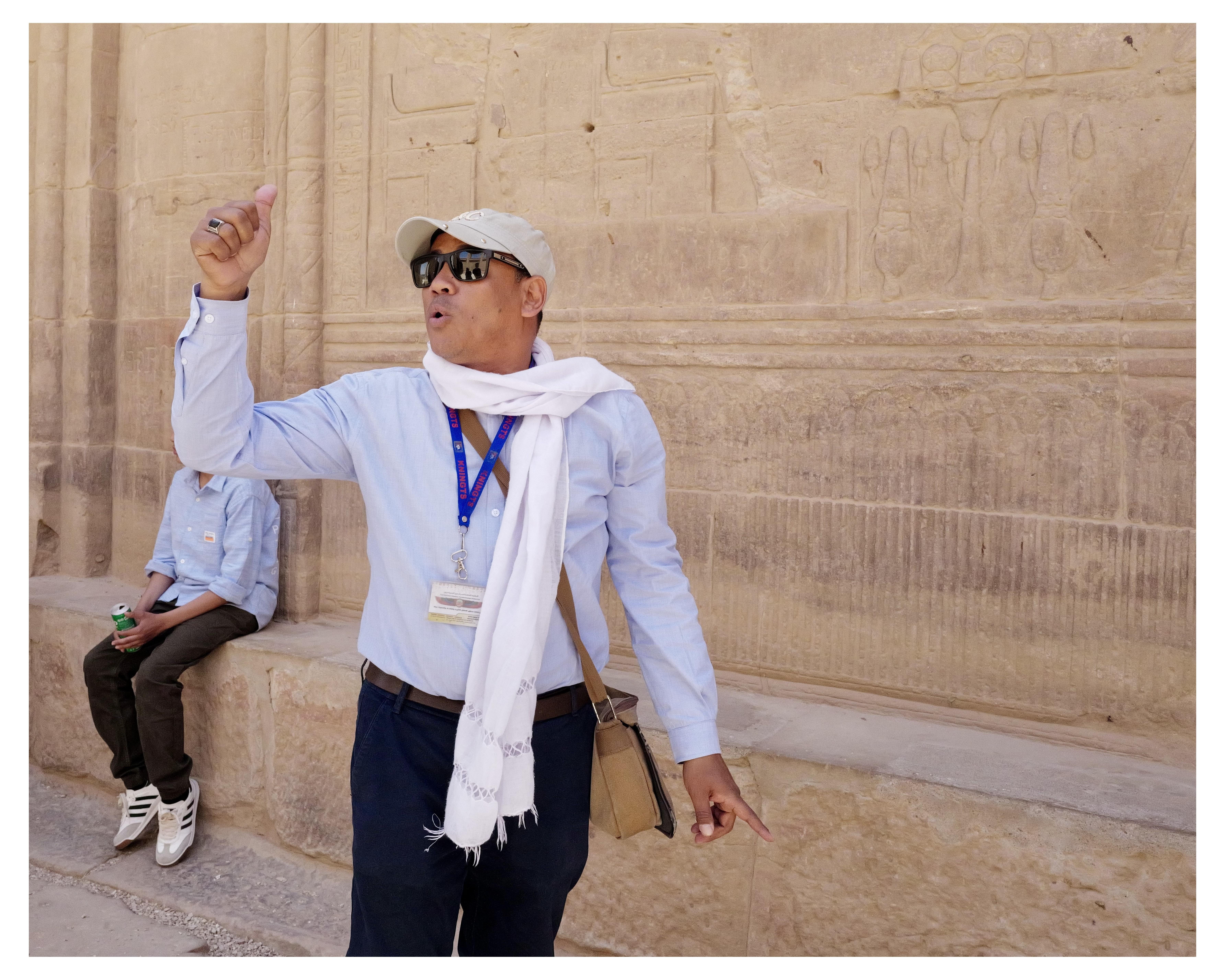 The image shows two people at what appears to be an archaeological or historical site. The main focus is a man wearing a white hat, sunglasses, and a light blue shirt, paired with a white scarf and dark pants. He is animatedly gesturing upwards, which indicates he might be explaining or describing something about the location. He also has a badge around his neck, suggesting he could be a tour guide.

The background features detailed carved hieroglyphs on a wall, indicating that this location could be an ancient Egyptian site. There's another individual sitting on a ledge in the background, perhaps listening to the guide.  The person’s face is obscured by the guide’s elbow.  This is Edfu temple in Egypt.