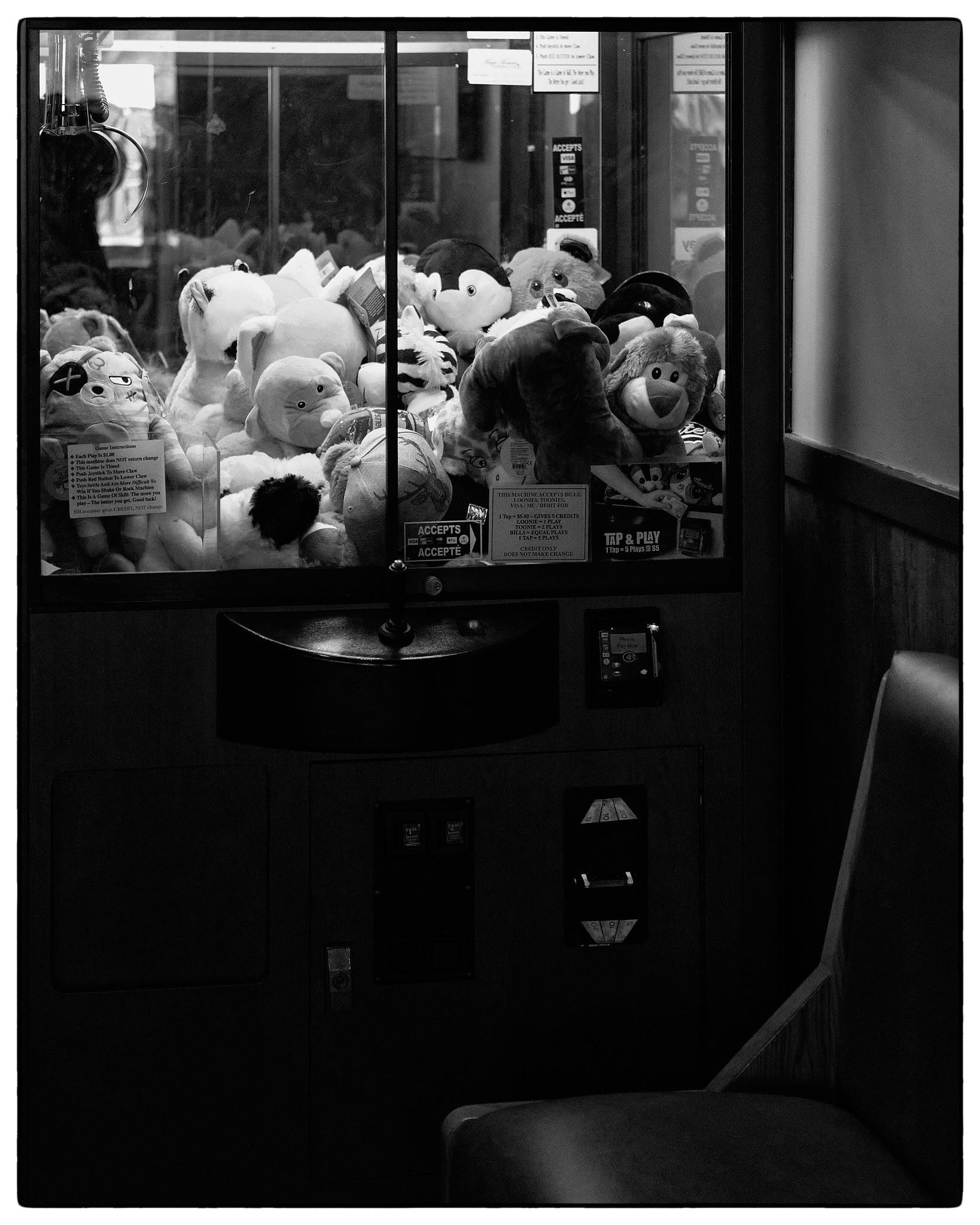 This black and white image features a claw machine filled with stuffed animals. The machine has glass windows through which various plush toys are visible, including what appear to be a monkey, a penguin, and a bear among others. To the right of the machine is a chair, suggesting that this setting might be in an indoor area where people can sit and play or watch others play. Additionally, there are visible labels on the machine indicating payment options and instructions for playing the game.