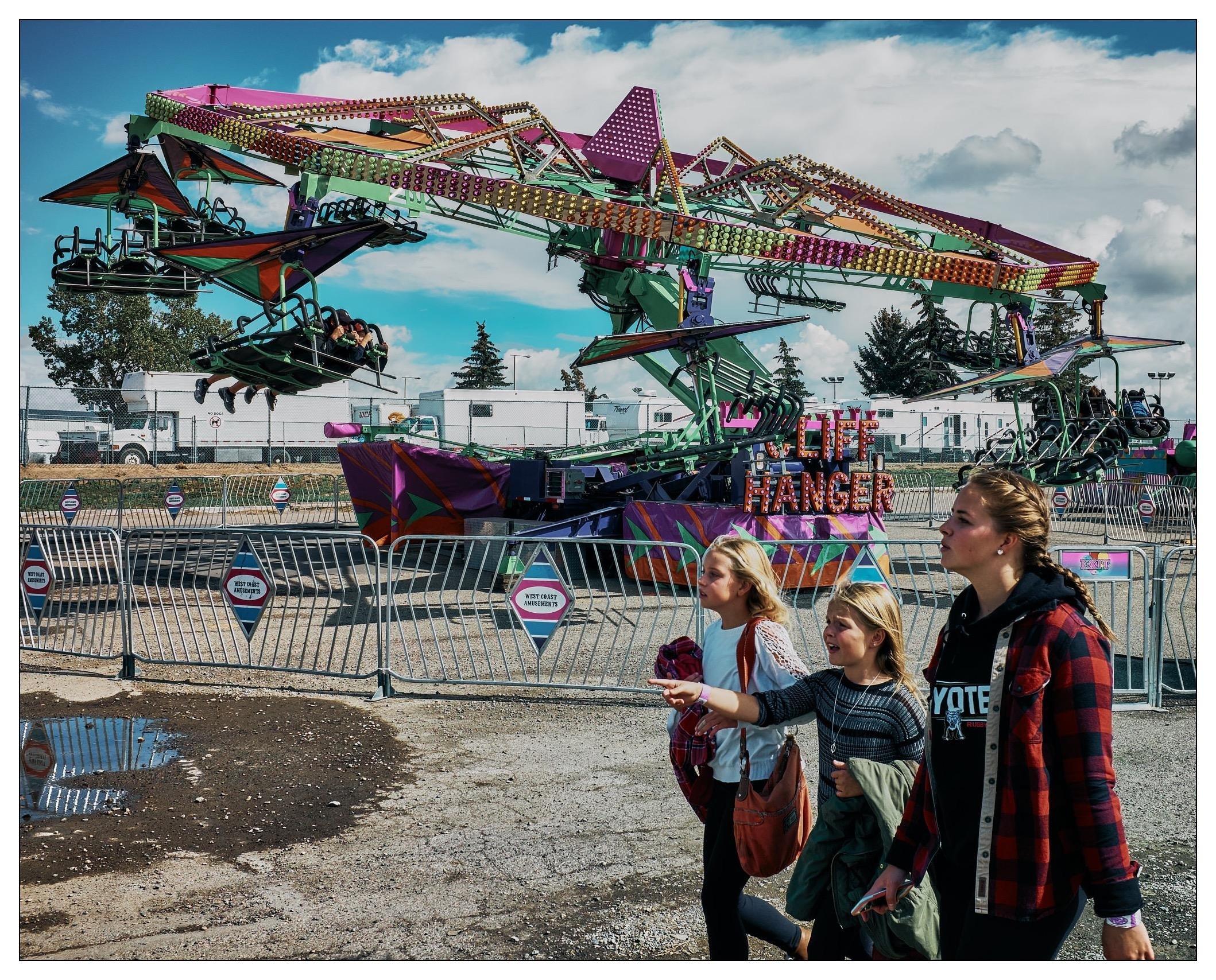 The image captures a vibrant scene at an amusement park or fair. There are three people, possibly family or friends, walking and looking toward a colorful amusement ride in motion, called "THE CLIFF HANGER." The ride is brightly decorated and people are strapped into it, lying flat as if they are flying. The setting includes a partly cloudy sky and there are visible puddles on the ground, suggesting recent rain. The ambience is lively, fitting the context of a fairground, which is further suggested by other installations and structures in the background, likely part of the fair setup.