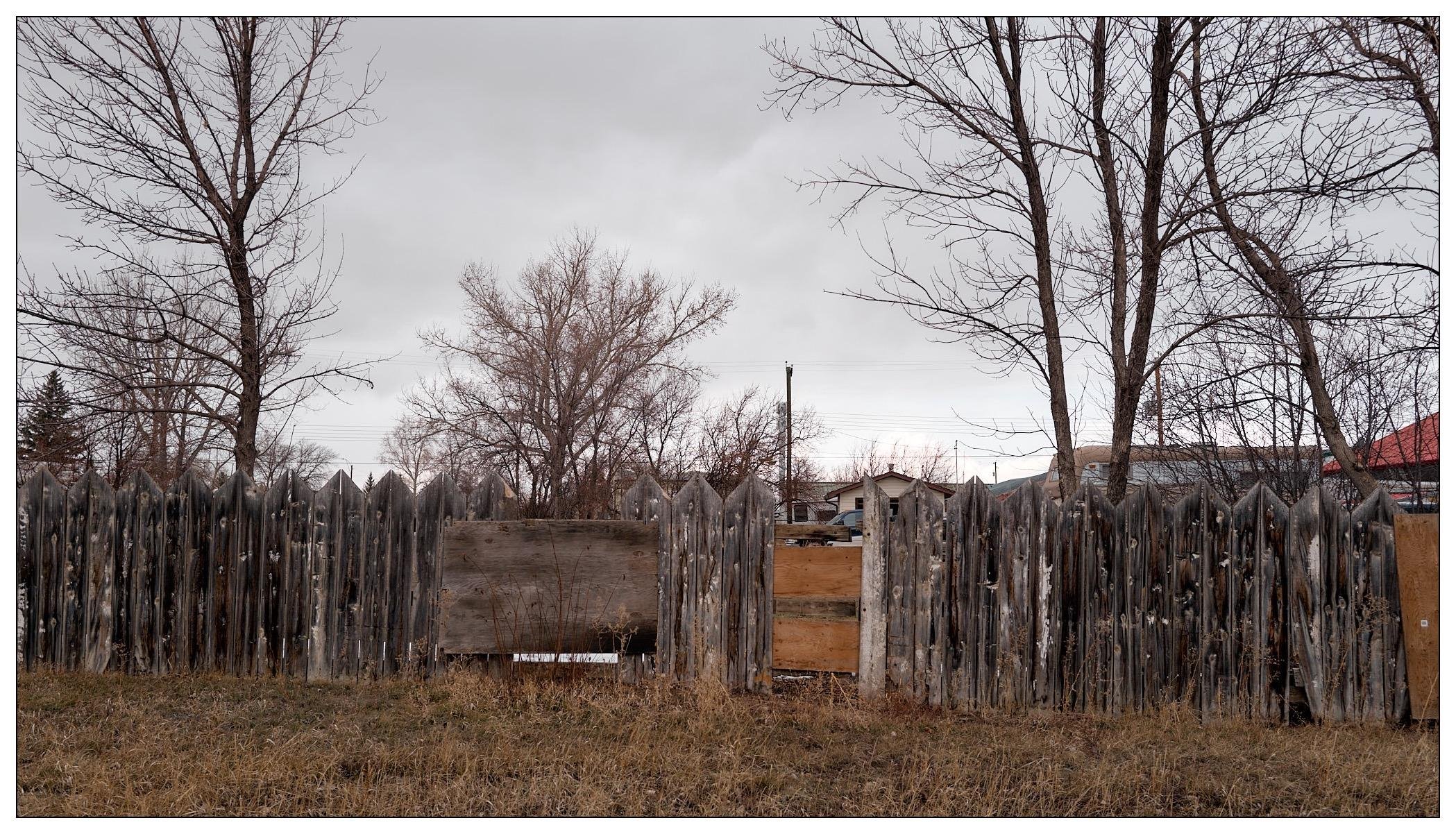 A weathered wooden picket fence in a rural or semi-rural setting. The fence, which appears aged and deteriorating, curves around from the left to the center of the frame. It features a makeshift repair made of plywood pieces. Behind the fence, bare trees and dry grass suggest it might be late fall or winter. The overall atmosphere is overcast, with a pale, cloudy sky. There are also hints of residential structures in the background beyond the trees and fence, indicating that this might be on the outskirts of a town or in a sparsely populated area.