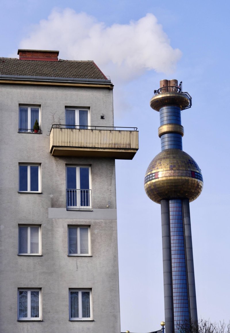 The image shows a contrast between an old residential building and a futuristic, artistic industrial tower. On the left, there is a gray apartment building with multiple windows and balconies, one of which has a small plant. The building appears weathered. On the right, a tall chimney with a unique design features a spherical, gold-tiled section and blue mosaic patterns, emitting white smoke into the sky. The scene highlights the coexistence of everyday urban life with striking industrial architecture.