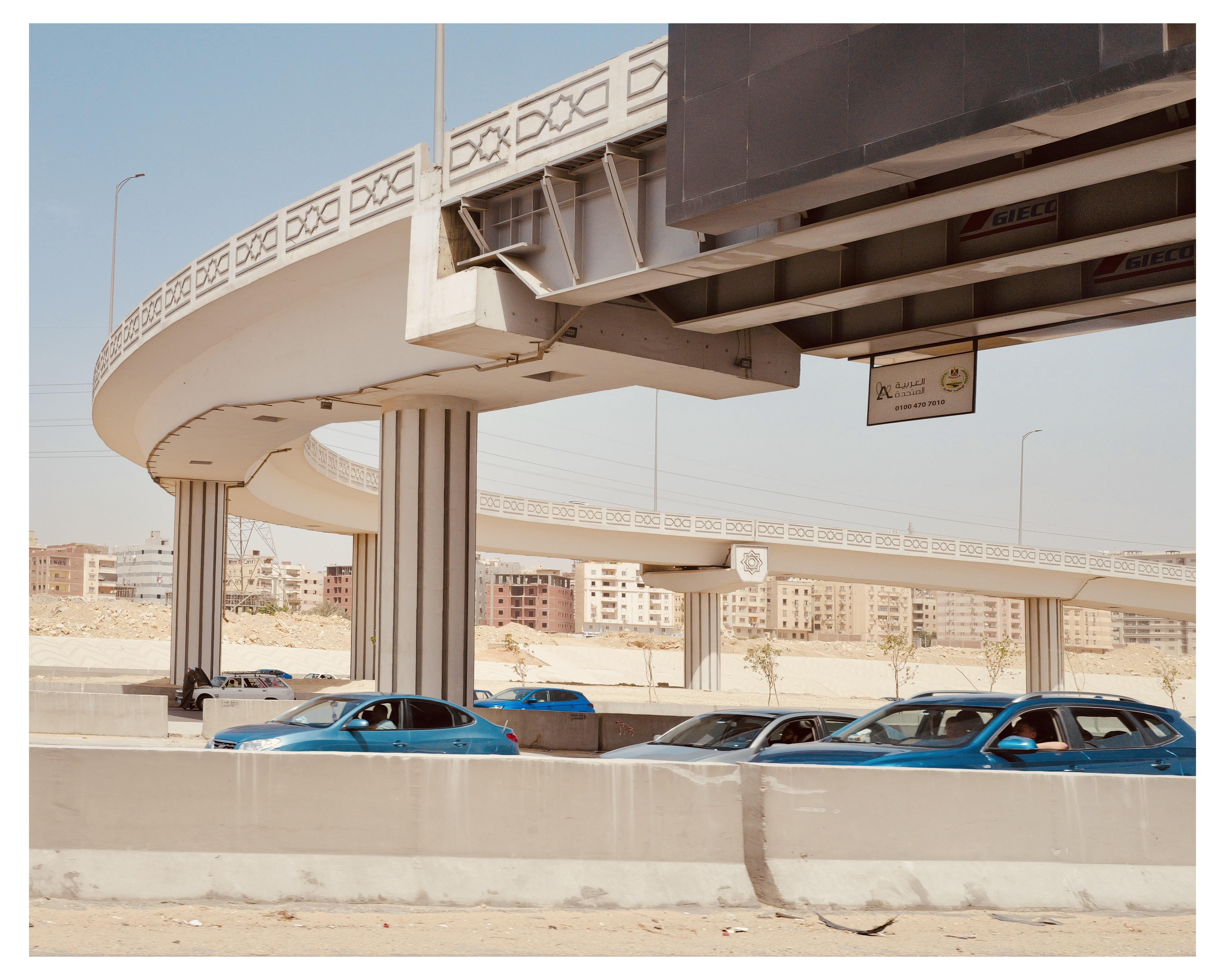 This image features a complex multi-level roadway with overhead highways and an exit ramp. The architecture includes several columns supporting the elevated roads. There are a number of cars, primarily in blue tones, traveling along and parked beside the road. In the background, there are buildings that appear to be part of an urban residential or commercial area. The scene is set against a clear sky, suggesting a sunny, calm day. The signage and structures' design might indicate that this location is in a region using Arabic script, possibly the Middle East.